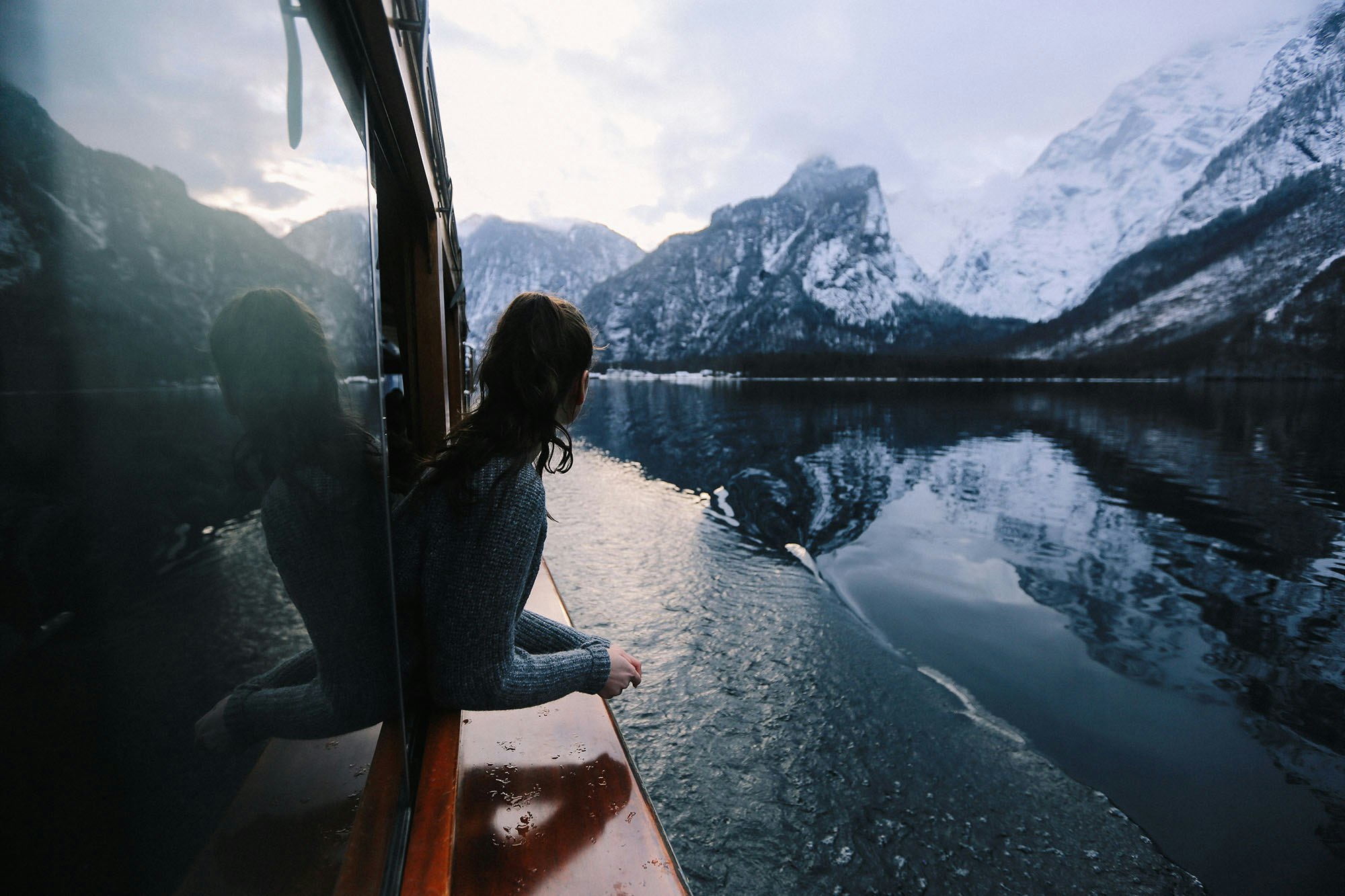 A woman leans out from the window of a small boat cruising through dark blue waters with snow-covered mountains in the distance.