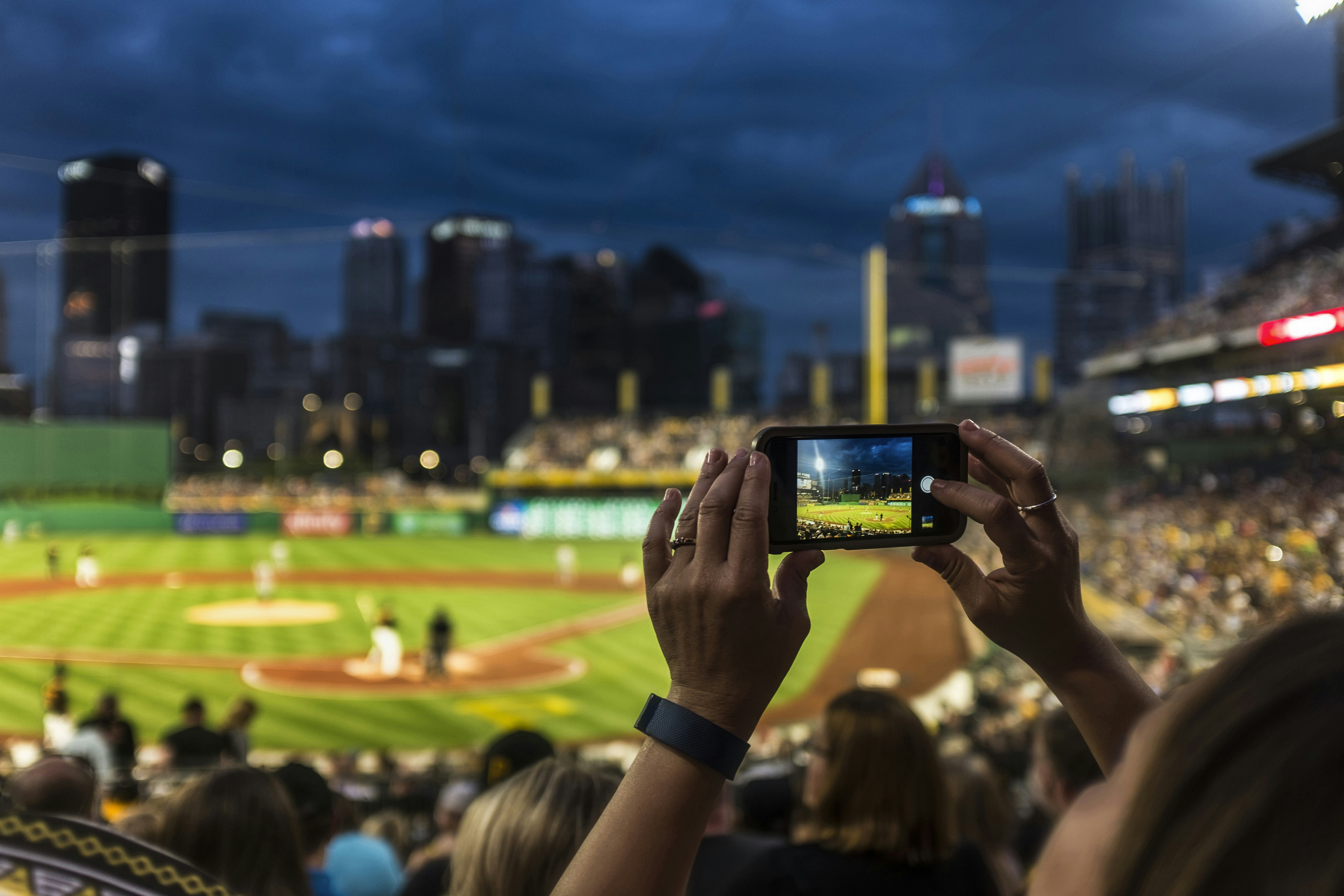 Inside a floodlit baseball stadium at night, a woman is holding up a cellphone and taking a photo. Her hands and the phone are in sharp focus, while the rest of the scene is blurred.