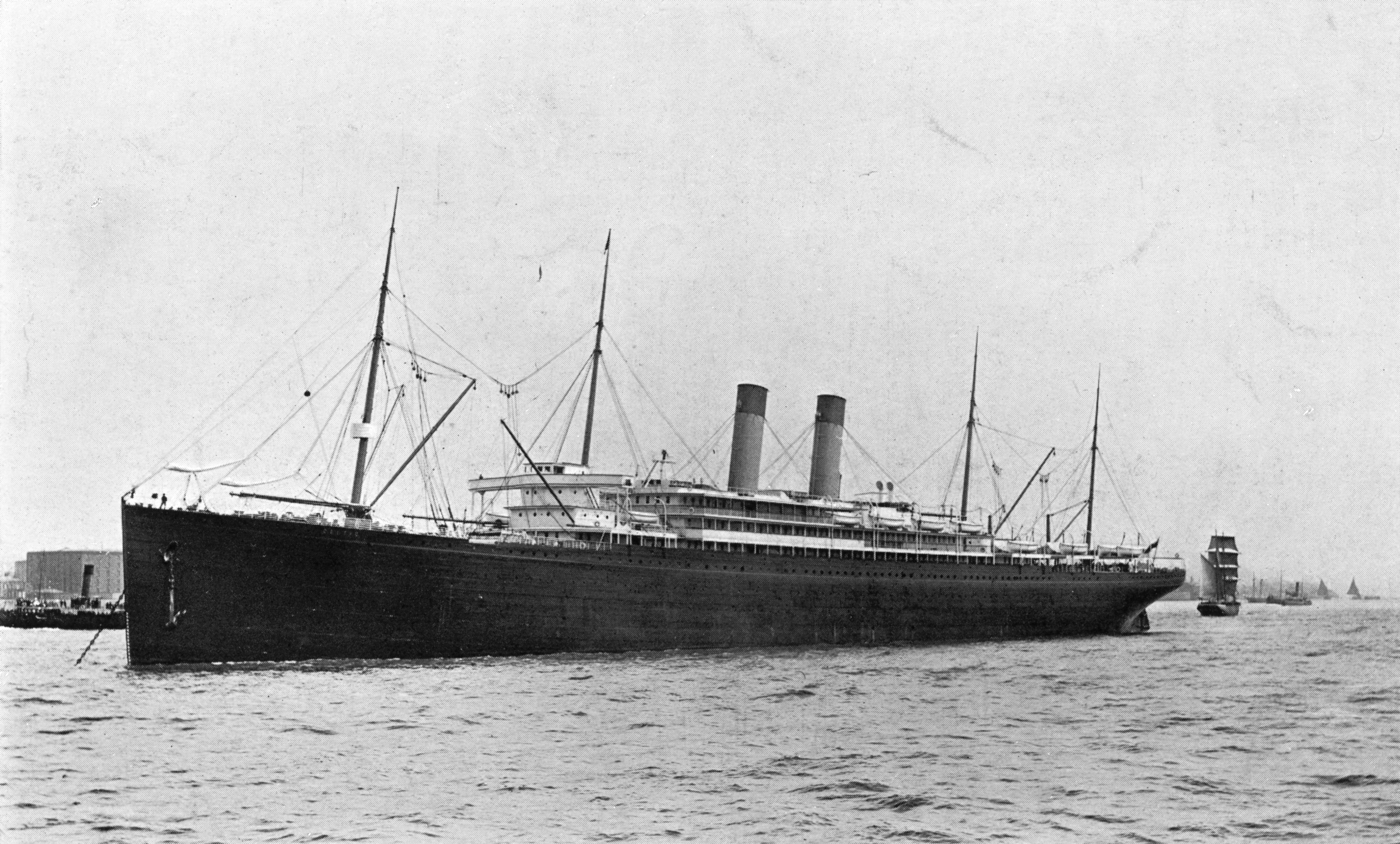 The RMS Celtic appears in this black and white photograph circa 1913 with two smoke stacks, four masts, and a long black hull angled towards the left side of the frame sailing towards, but past the viewer.