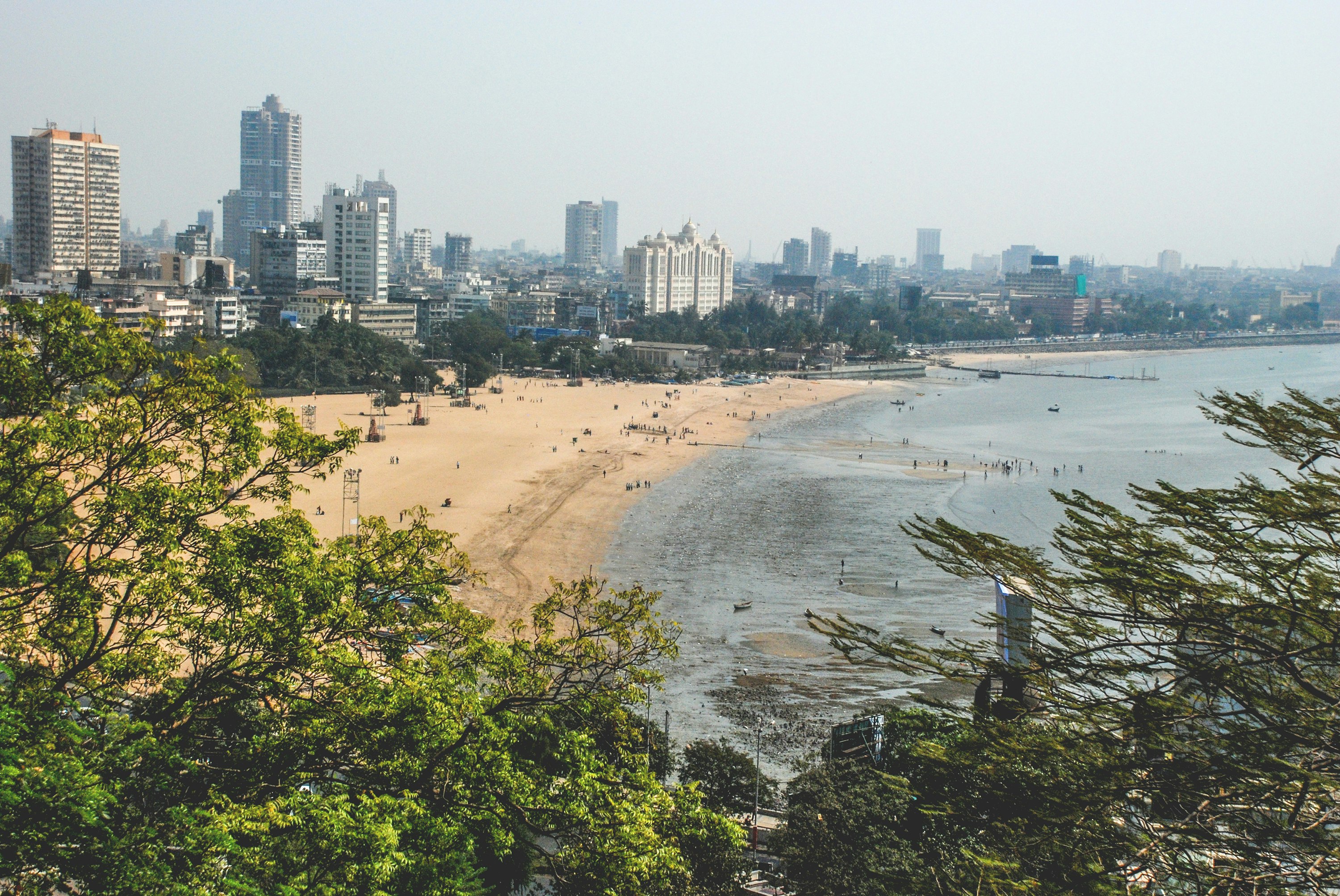 A view overlooking Juhu Beach with the Mumbai cityscape in the background. The photo is framed by green foliage. Crowds of people are visible on the wide slab of sand.