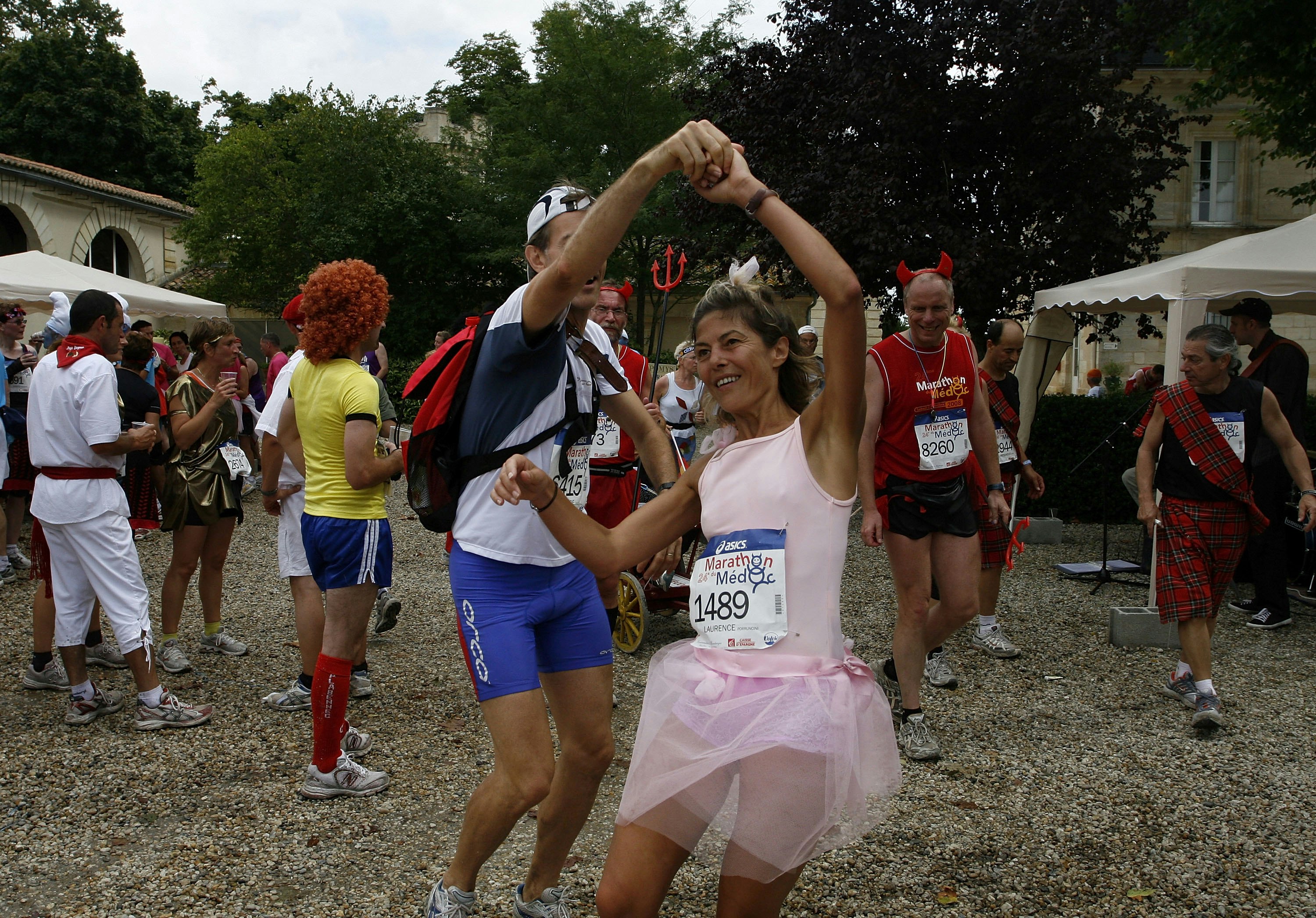 Several marathon participants are milling around at a vineyard wine-tasting station. In the foreground, a man and a woman (who is dressed as a ballerina) are dancing.