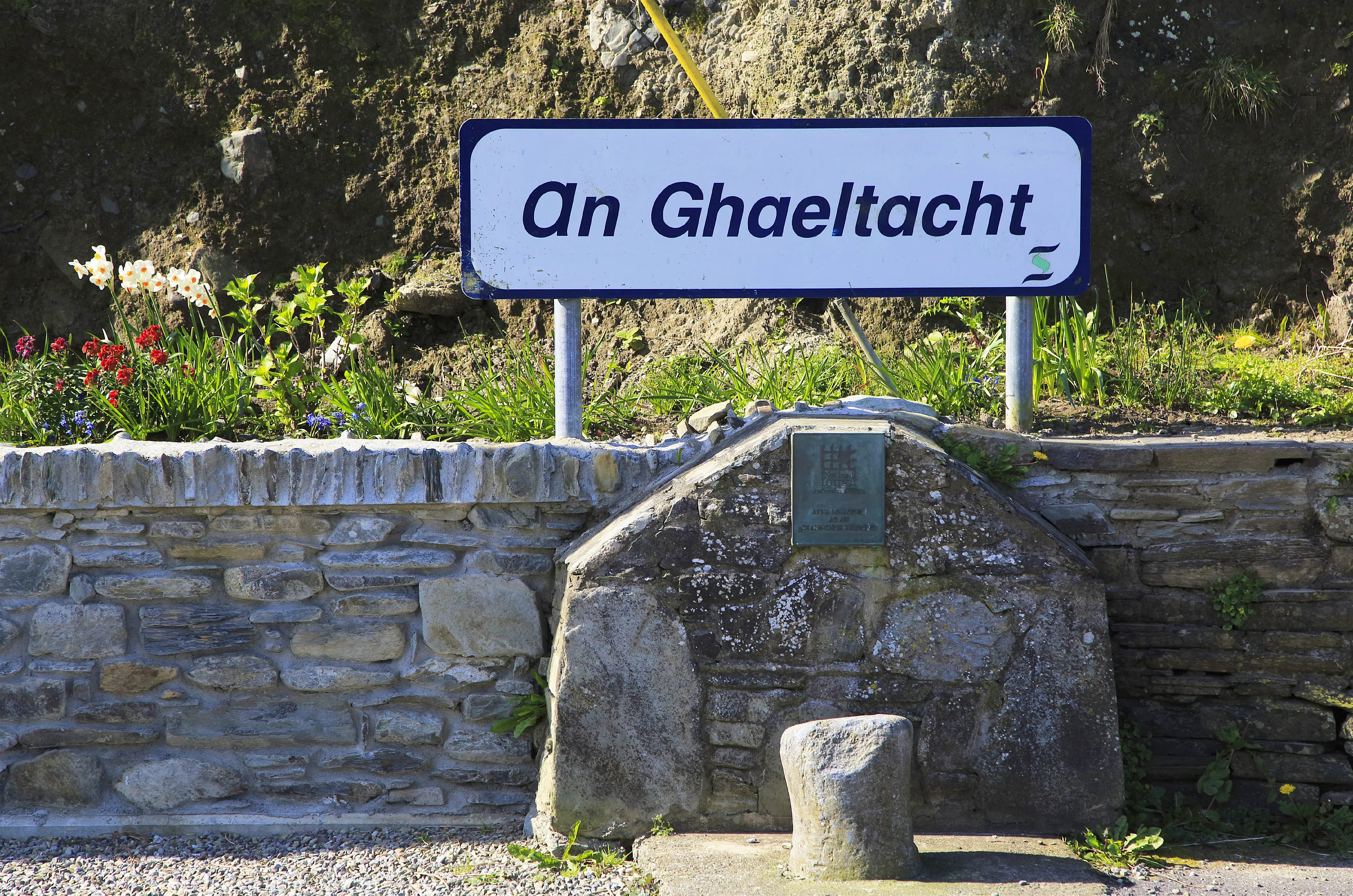 ghaeltacht-cape-clear-ireland.jpg A white rectangular sign with rounded edges reads "an ghaeltacht" in a blue italicized sans-serif font. It sits on a long low stone wall with a small illegible plaque just below the white sign. It indicates this is a place where Gaeilge is still the primary language