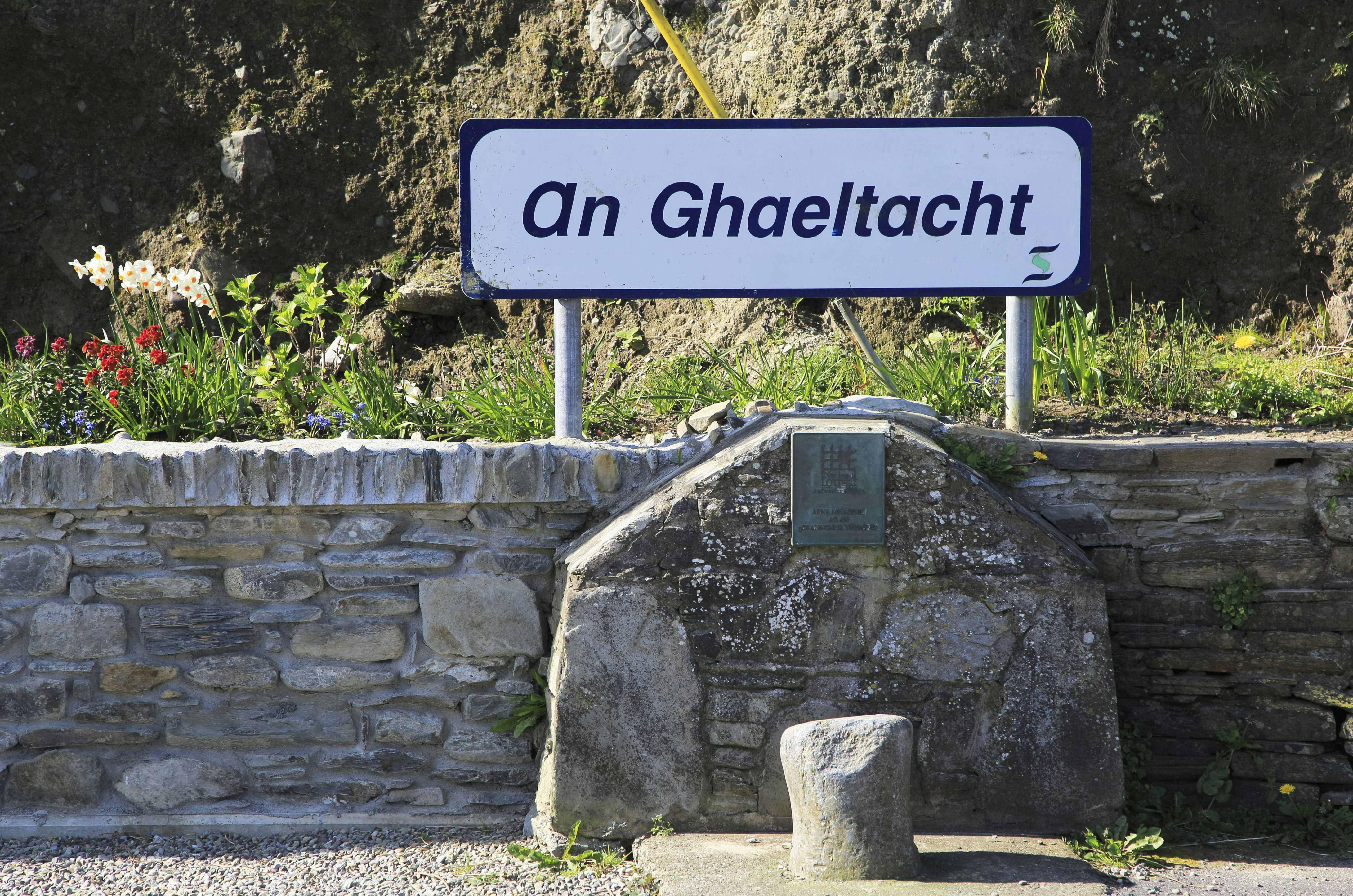 A white rectangular sign with rounded edges reads "an ghaeltacht"  in a blue italicized sans-serif font. It sits on a long low stone wall with a small illegible plaque just below the white sign. It indicates this is a place where Gaeilge is still the primary language