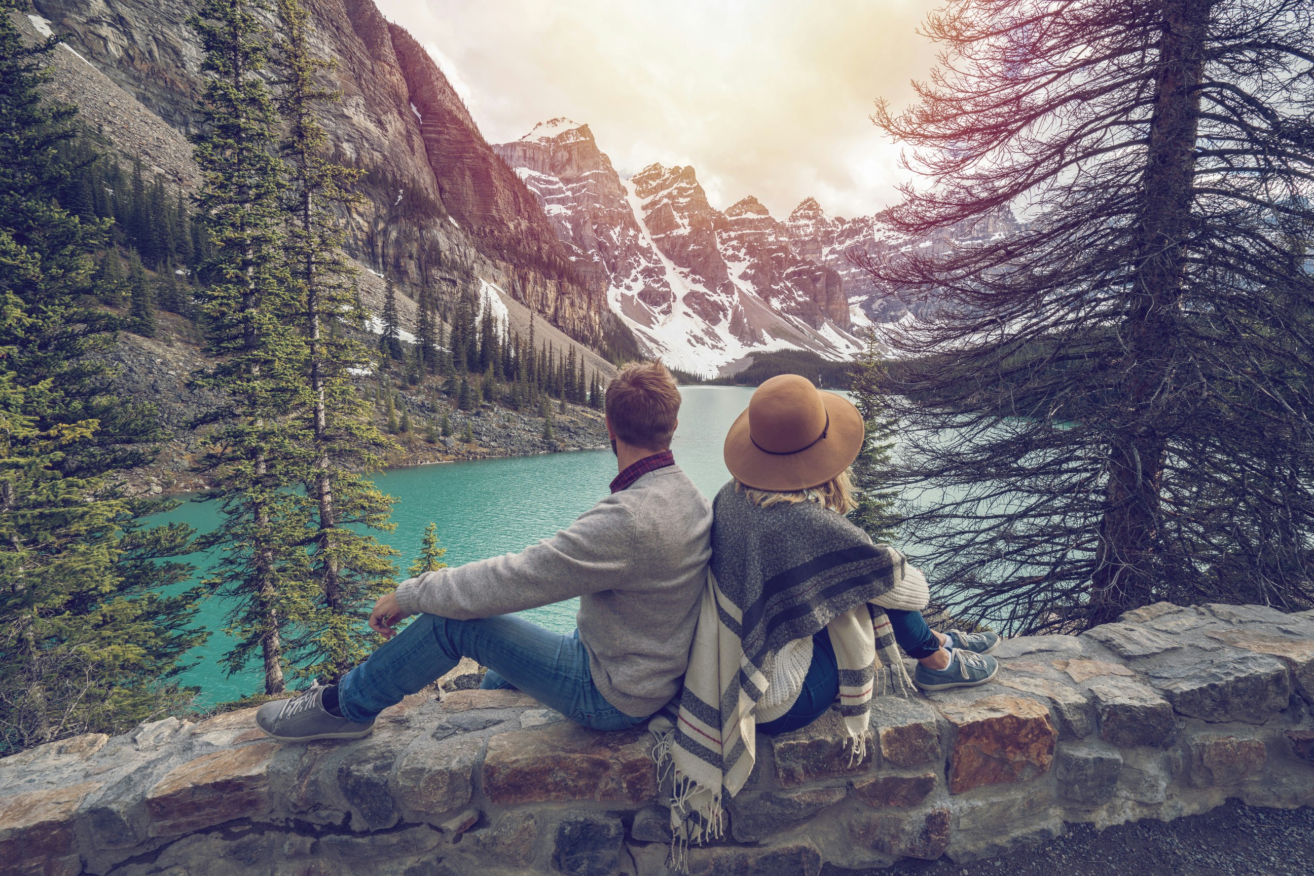 A couple sit back to back on a low stone wall. They are looking out towards a lake, beyond which are huge mountains laced with snow. There are tall pine trees in the foreground.
