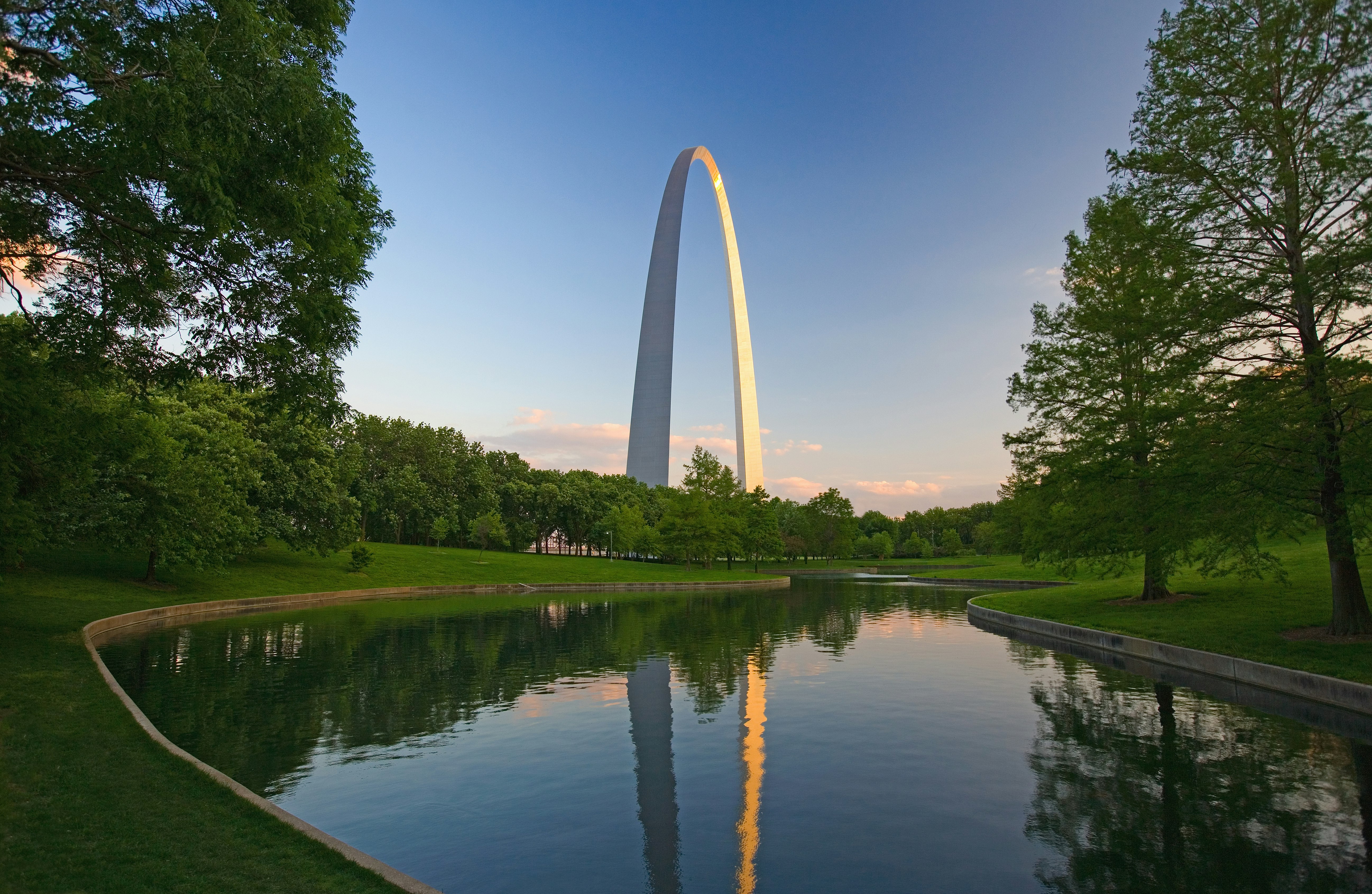 The silver expanse of the Gateway Arch is seen almost in profile, the closest end catching the bright warmth of the afternoon sun. The foreground of the image is dominated by a blue body of water flanked by a bright green lawn and green, shady trees.