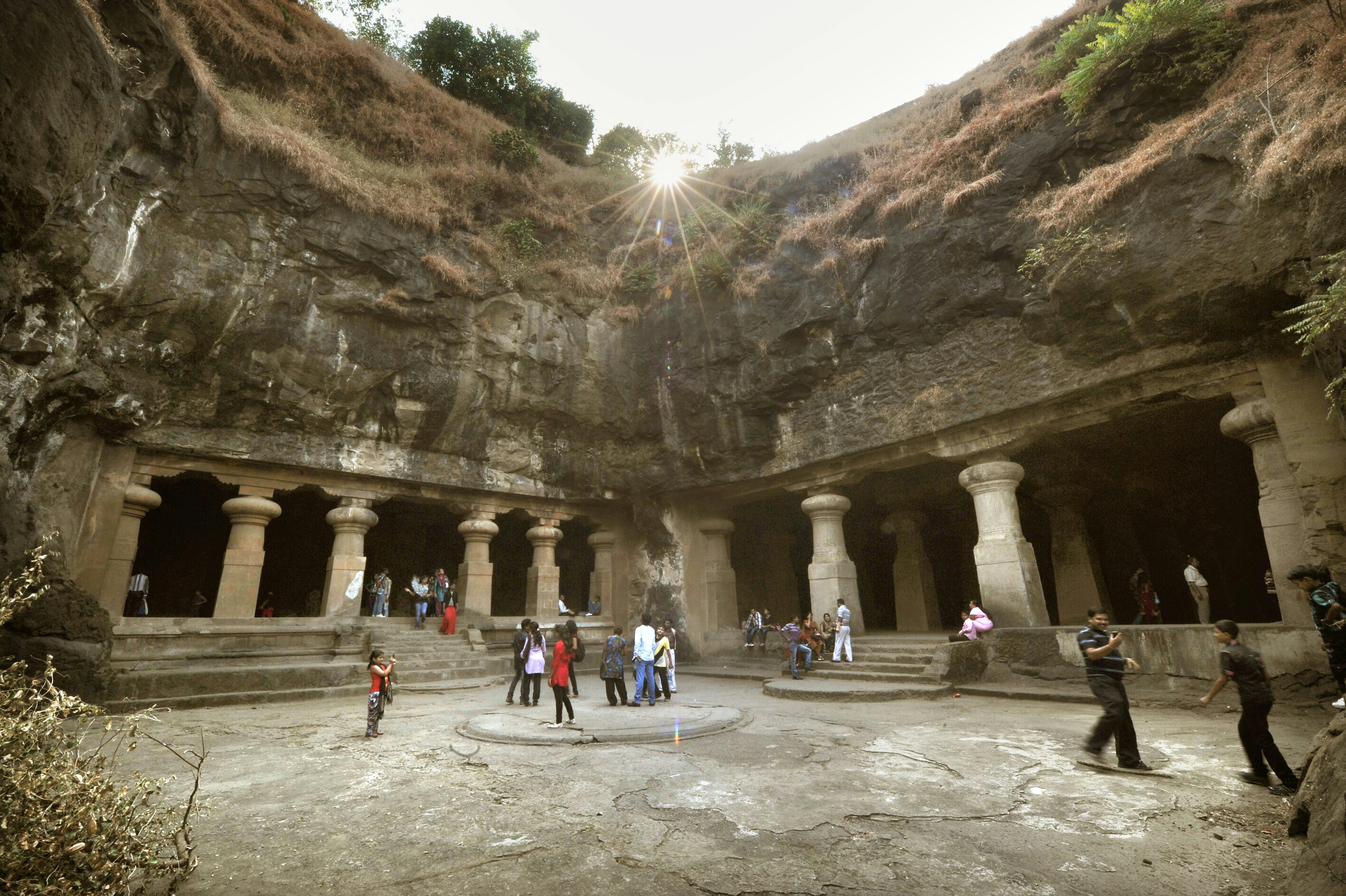 Groups of visitors wander around the Elephanta Cave complex in Mumbai. The complex includes several open-air 'courtyards', as shown here, with the sunlight shining down from above.