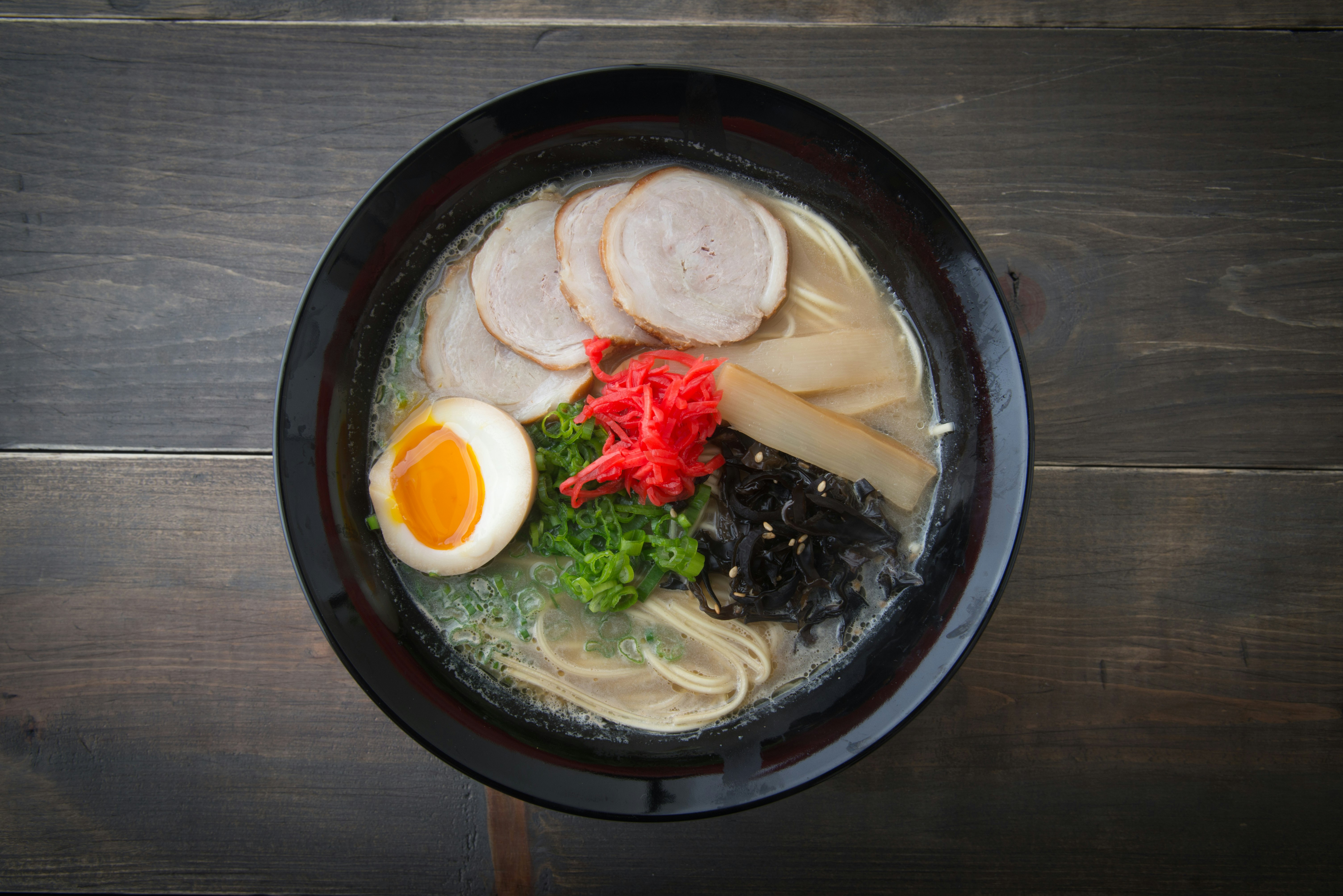 A glossy black bowl filled with ramen noodles, clear broth, round slices of pork, spring onions, half a soft-boiled egg, and other colourful ingredients.