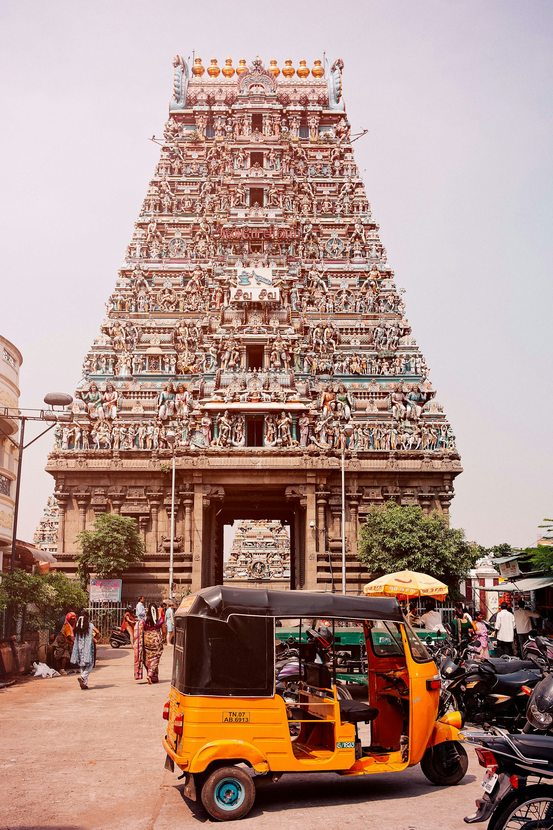 A bright orange motorized rickshaw with a black roof is parked in front of the colorful facade of the the Kapaleeswarar Temple in Chennai, which has several tiers like a pyramid covered in sculptures of humans and animals.