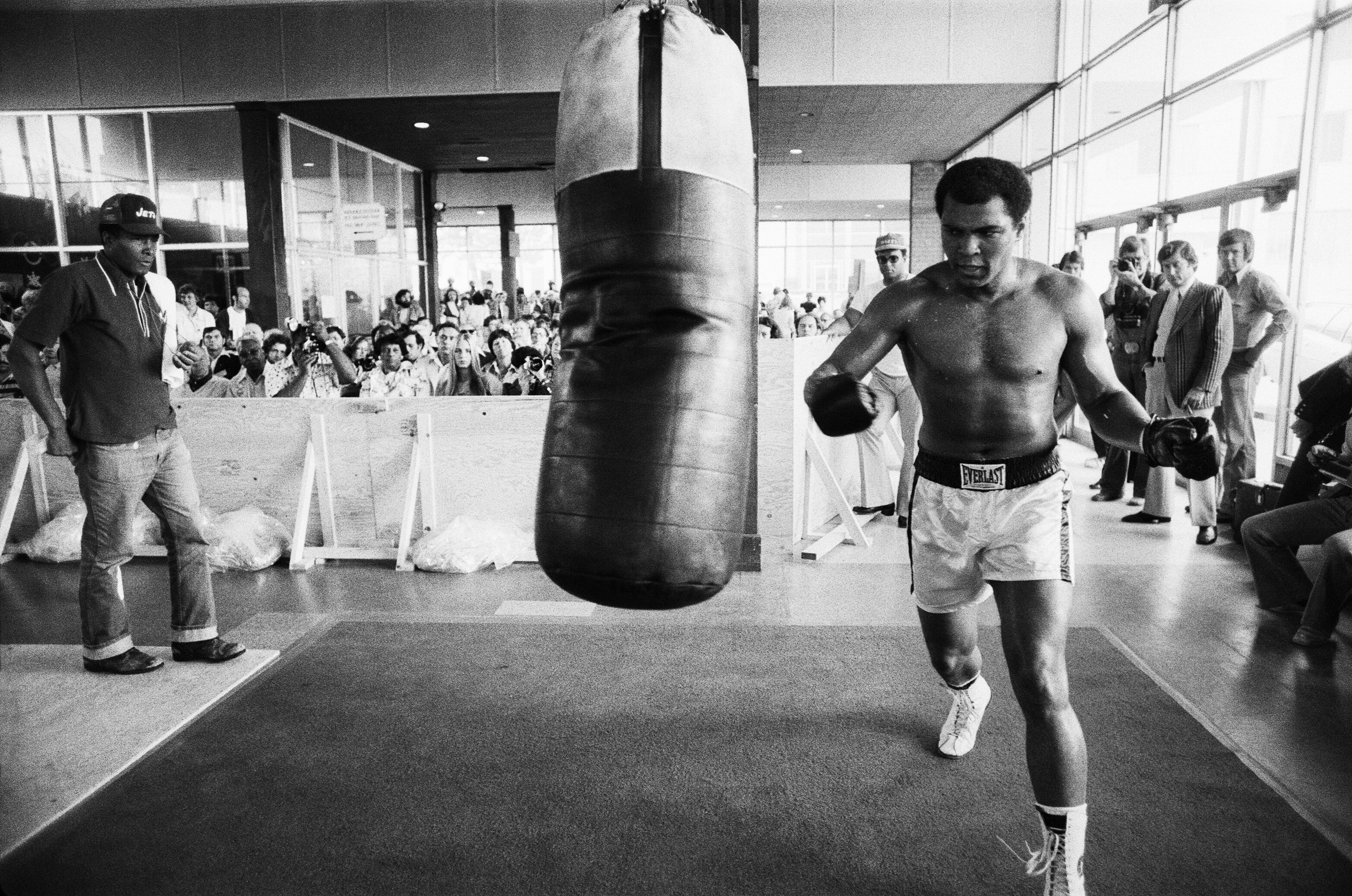 Muhammed Ali trains on a large punching bag at the Concord Hotel in Catskill Mountains with a seated crowd watching from behind a protective wooden barrier.