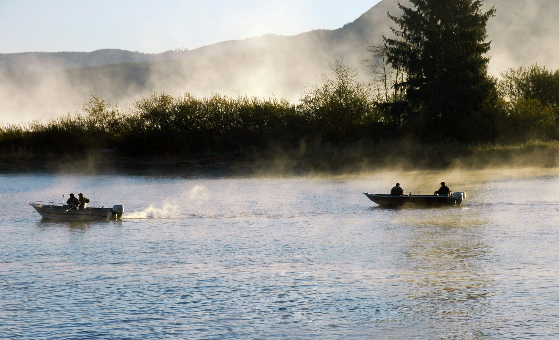 Two pairs of fishermen, each in small flat-bottomed motorboats, perch on the misty Nehalem River in Oregon. The water is flat and blue, barely ruffled by the breeze. In the background, shrubs and one large evergreen tree form dramatic silhouettes against a plume of fog that just obscures the blue mountains in the distance. 