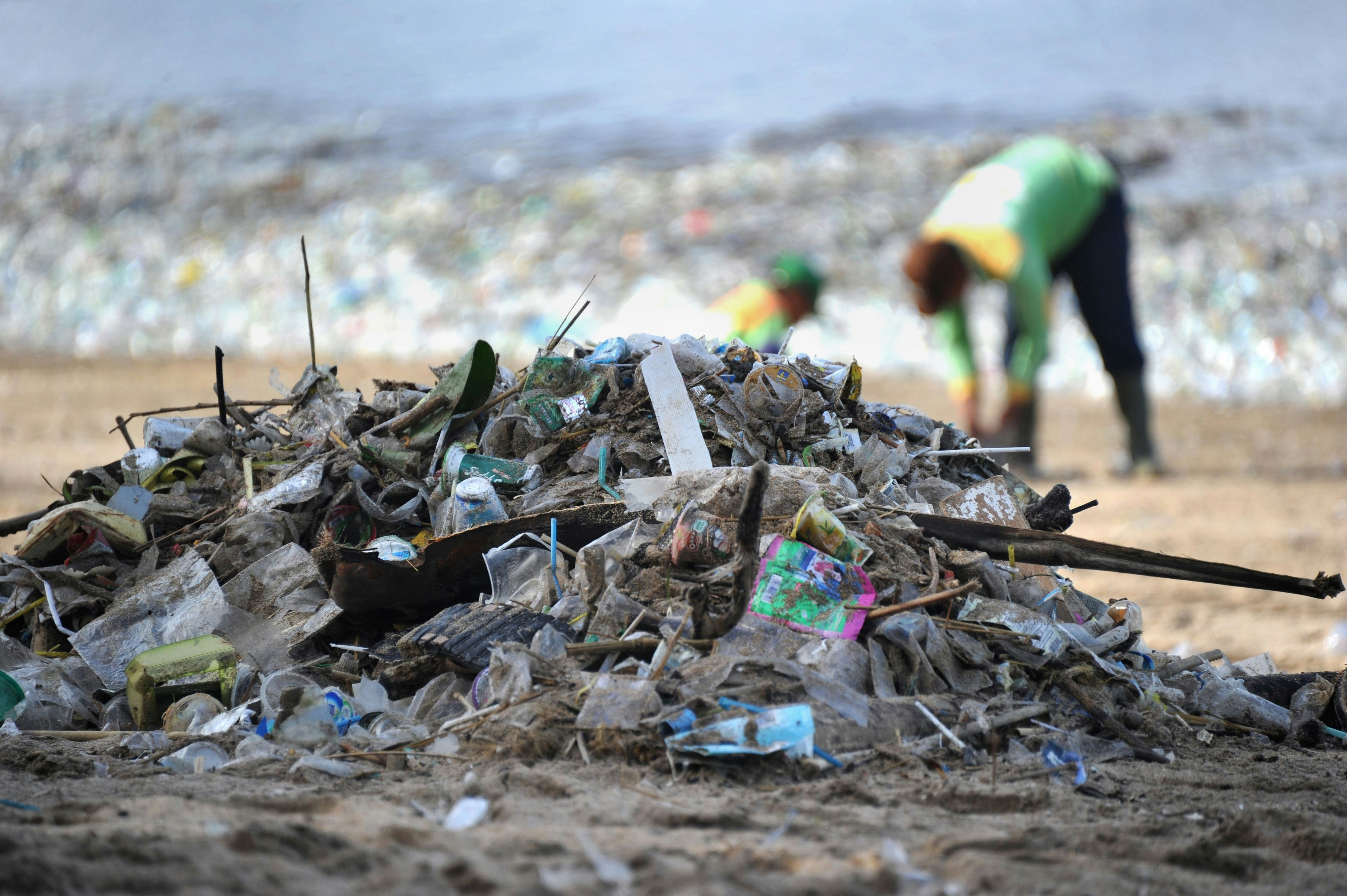 A mound of garbage on Kuta Beach in Bali. In the distance, and slightly out of focus, two people work to clear the litter from the sand.