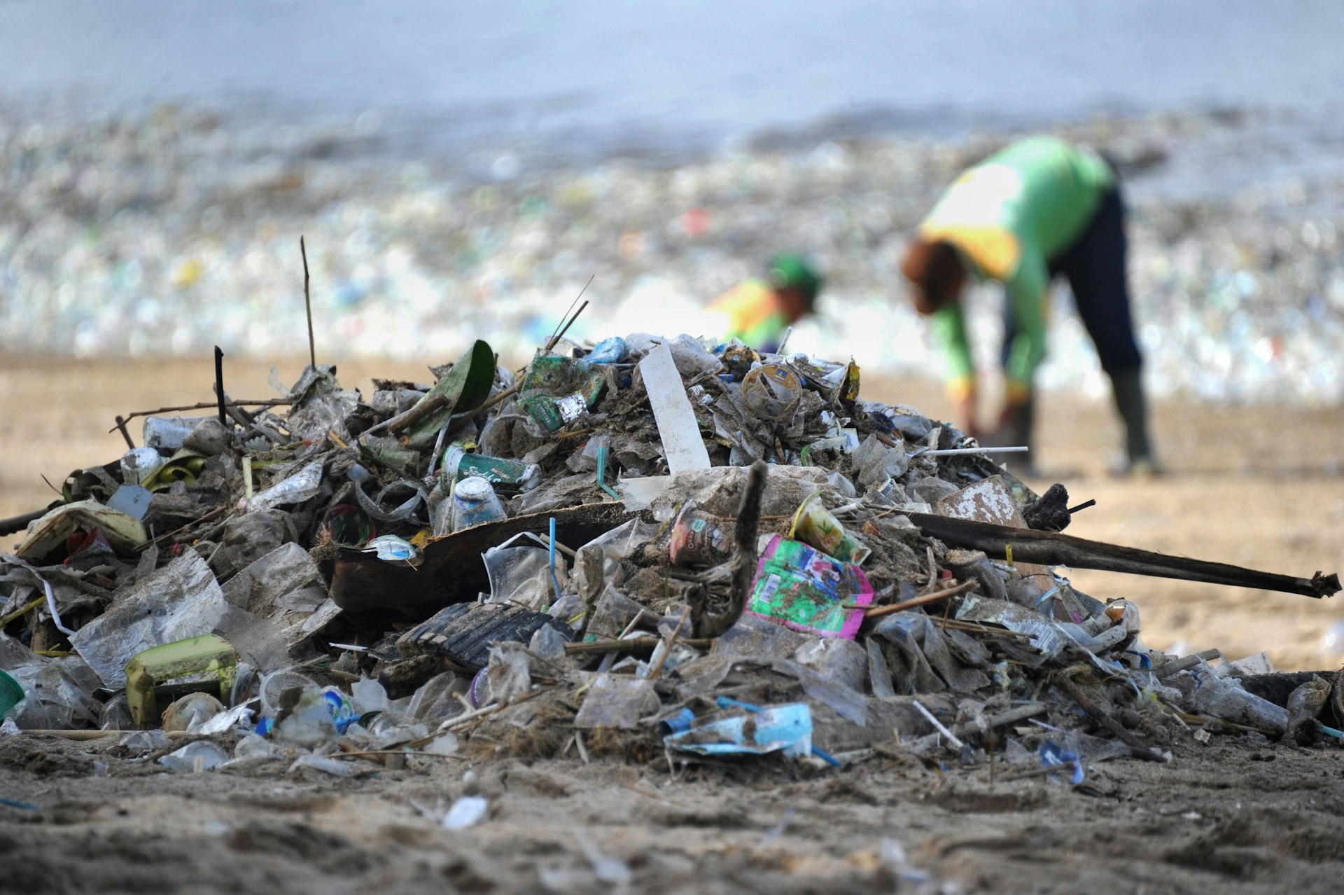 A mound of garbage on Kuta Beach in Bali. In the distance, and slightly out of focus, two people work to clear the litter from the sand.