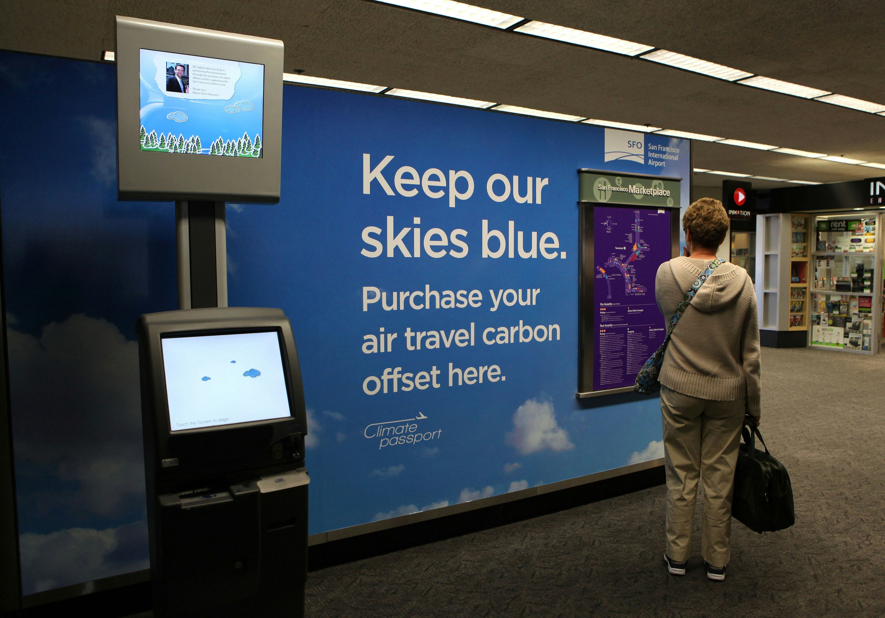 A female traveler with short blonde hair in khaki slacks and a tan sweater stands next to a carbon offset kiosk in the San Francisco International Airport. A large sign on a blue background with clouds reads "Keep our skies blue. Purchase your air travel carbon offset here" in a white san serif font  