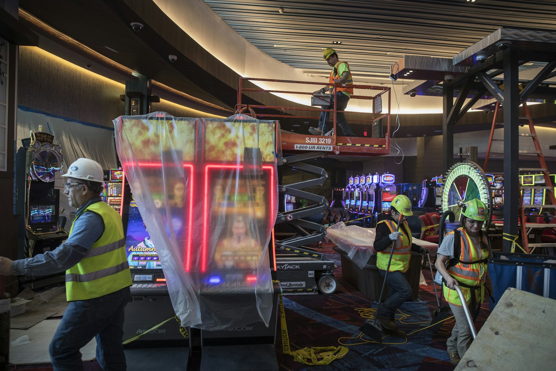 Construction workers in bright yellow safety vests and white hard hats work amidst brand new casino slot machines, roulette wheels, and other electronic games still wrapped in plastic. One holds a broom, one is vacuuming, and another stands on a scissor lift. A fourth on the left frame is gesturing to something just out of sight.