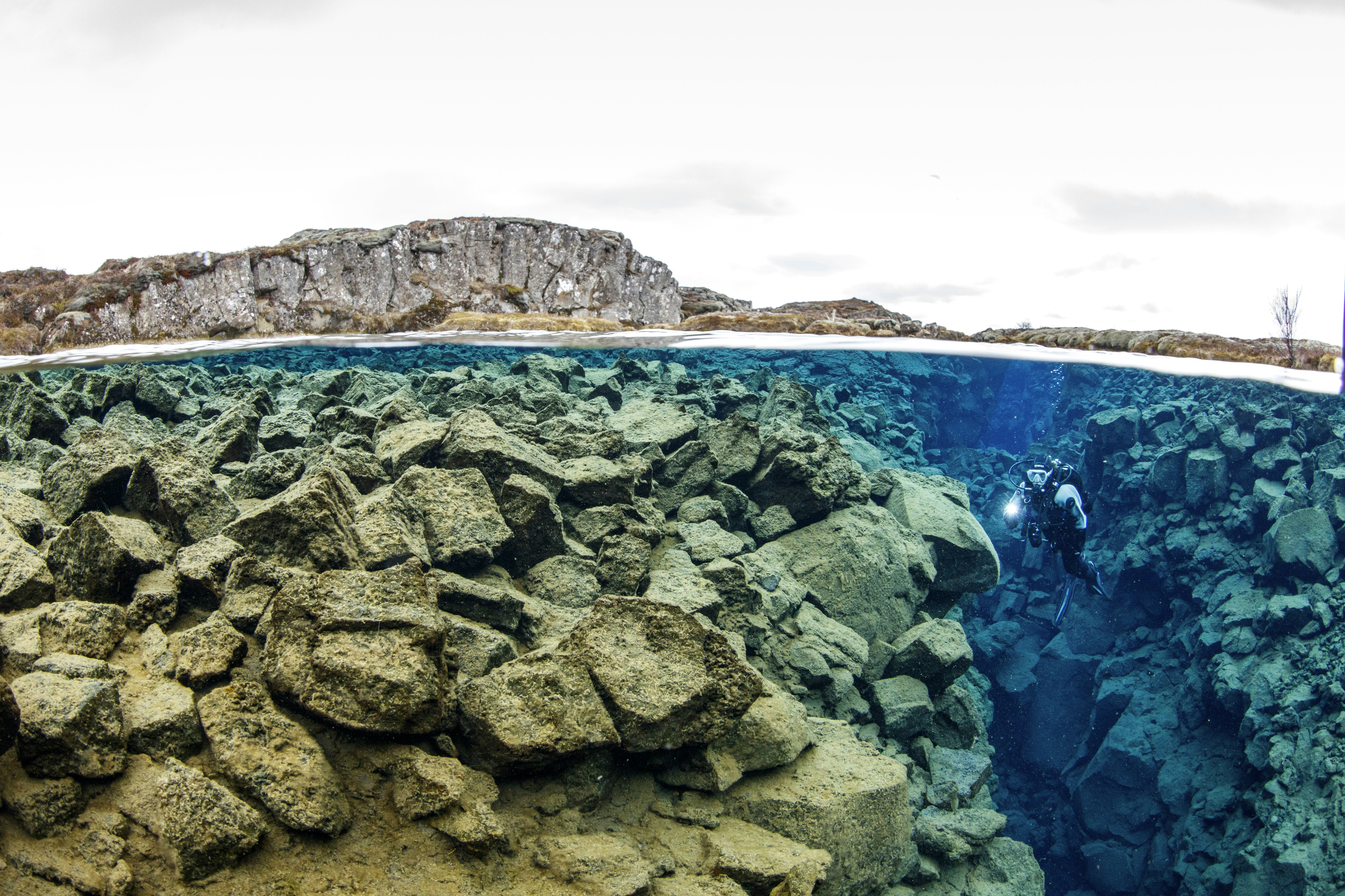 The top third of the shot shows the grey, rocky shore around the Silfra Fissure in Thingvellir National Park, while the bottom two thirds show what's under water, with a gradient of the tan, grey, turquoise, and deep lbue waters that are perfectly clear, and show a diver with a white light between two tectonic plates that form a narrow, rocky canyon