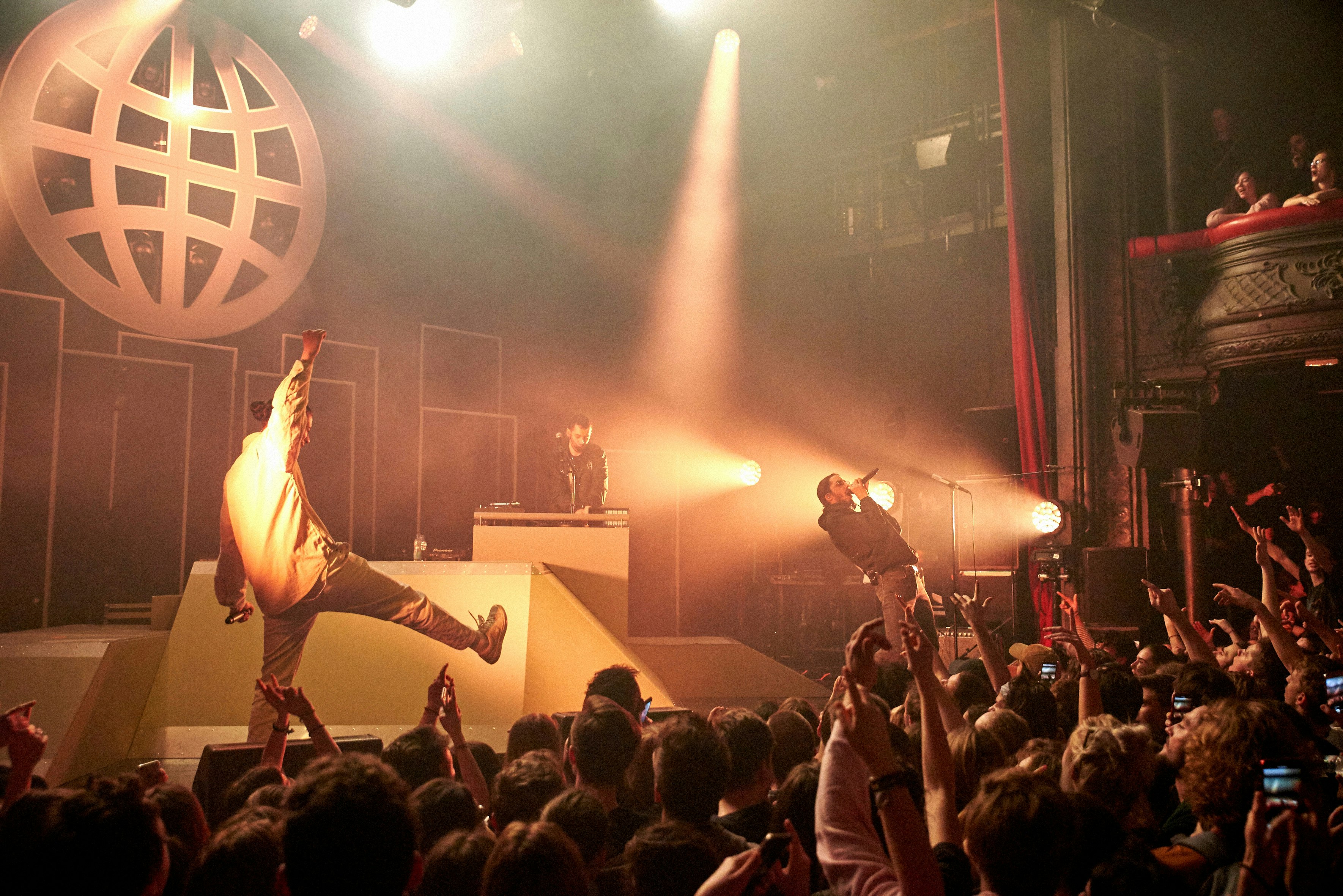 A rap group performs on the stage of La Cigale in Paris. An animated crowd, in shadow beneath the stage, watch on.