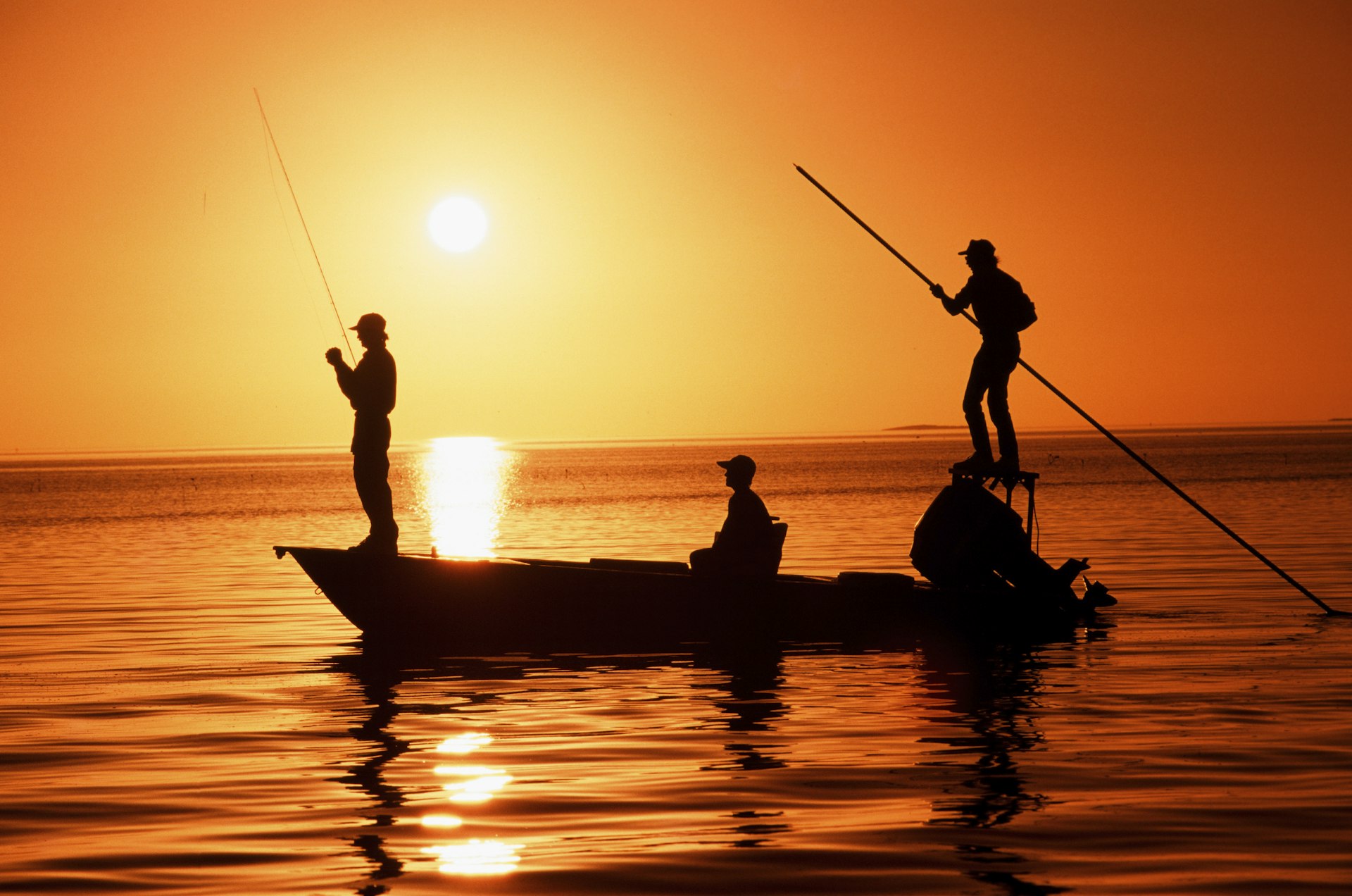 Three people are silhouettes against a vivid sunset as they stand and sit in a rowing boat. One of them holds a fishing rod