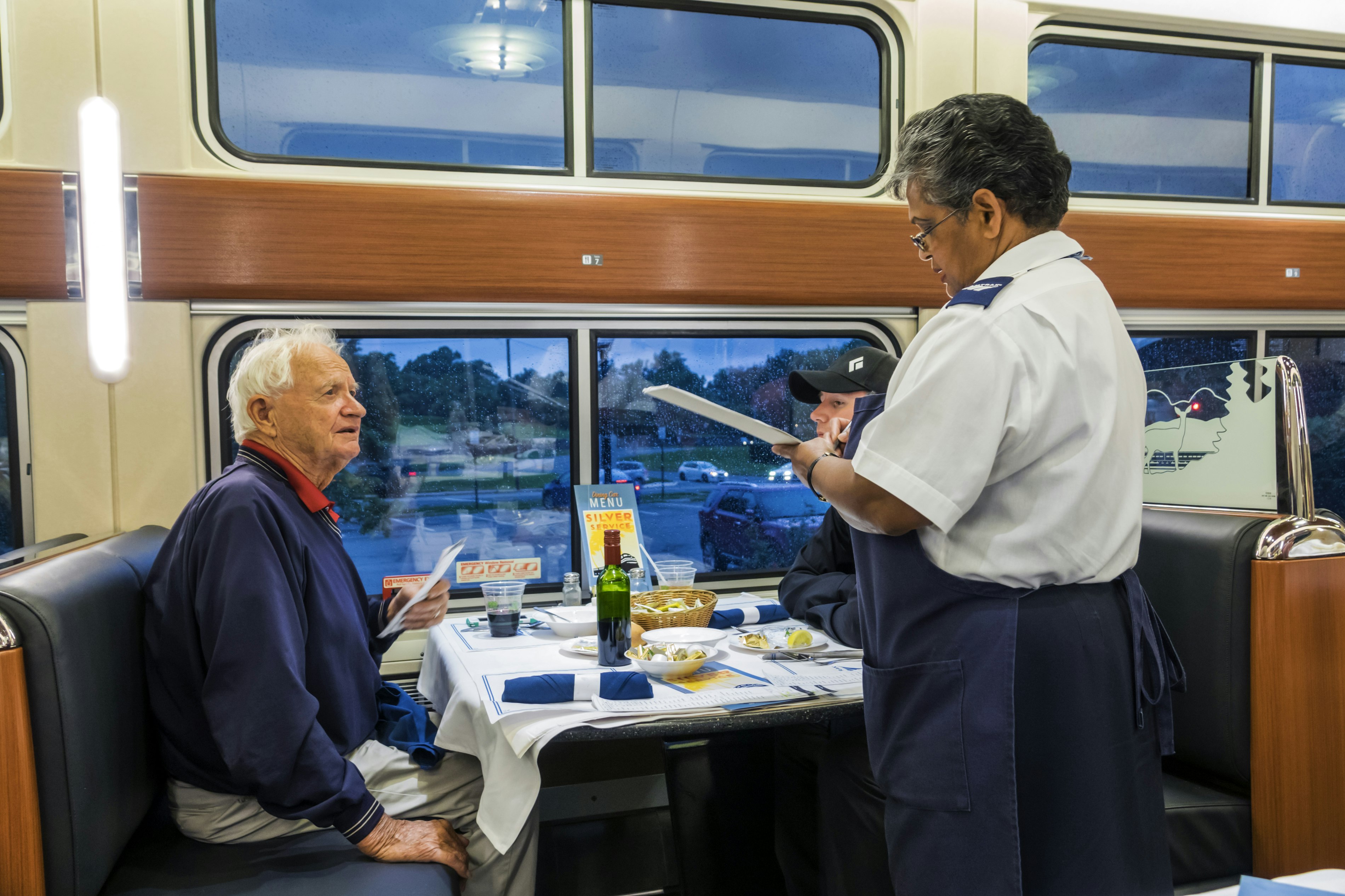 A waitress in a white tailored shirt with Amtrak-branded epaulets and a blue apron takes an order from a senior gentleman with white hair and a blue sweatshirt in the traditional Amtrak dining car, complete with white table cloths, blue cloth napkins, a green wine bottle, wicker bread basket, and a view of a train station parking lot at twilight.