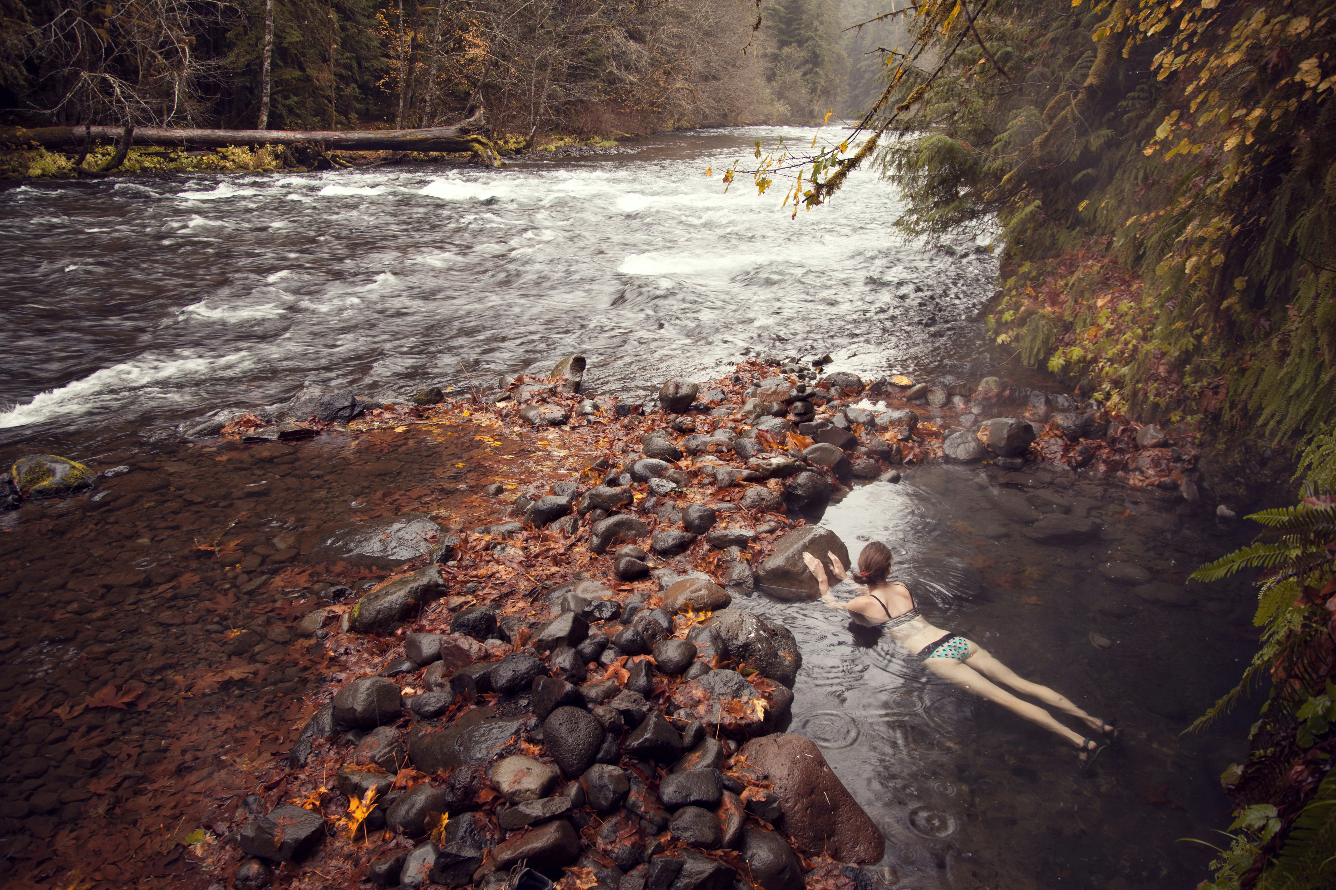 A young, thin woman in a green and white polka dotted bikini with a black waistband lays on her stomach in an Oregon hot spring overlooking a river just a few yards away. All around the spring are river rocks and leaves in autumnal colors, contrasting with the grey tint of the water except for whitecaps on the river rapids.
