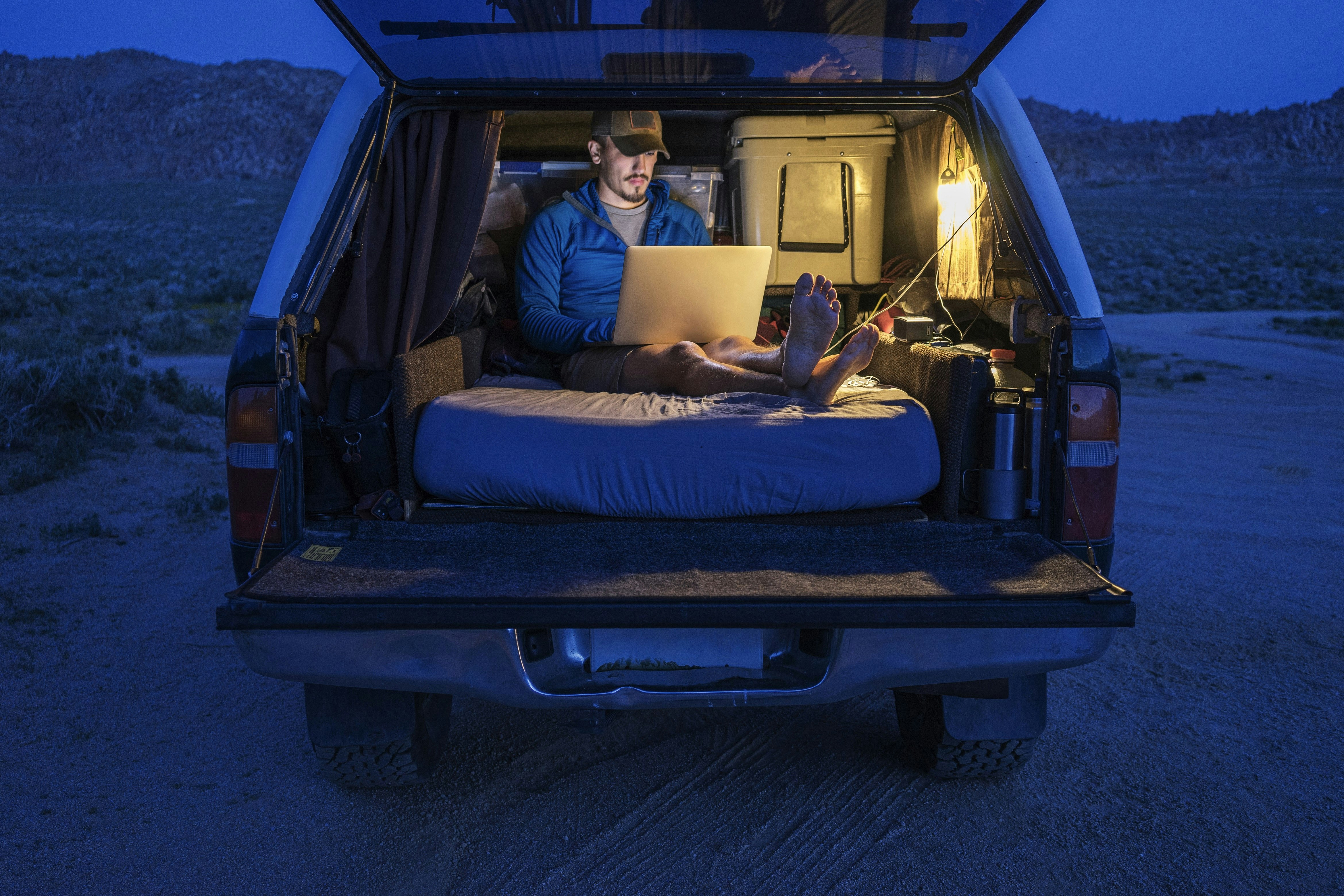 A man works on a laptop while sitting on a mattress in the back of a car at night time.