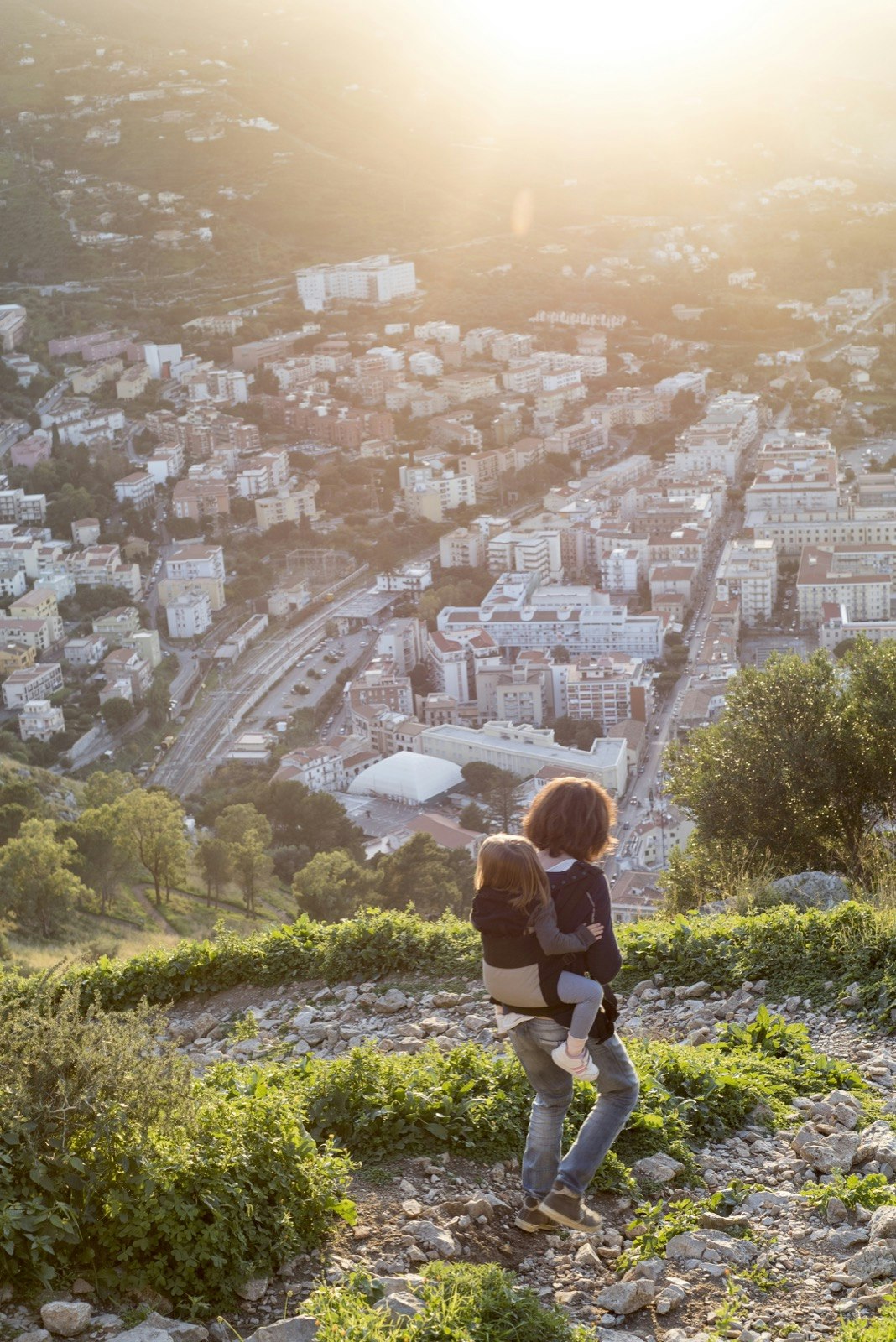 A woman with a child in her backpack walks down a steep path towards the city of Cefalu in Sicily during a sunset on a trip to Sicily with toddlers in tow