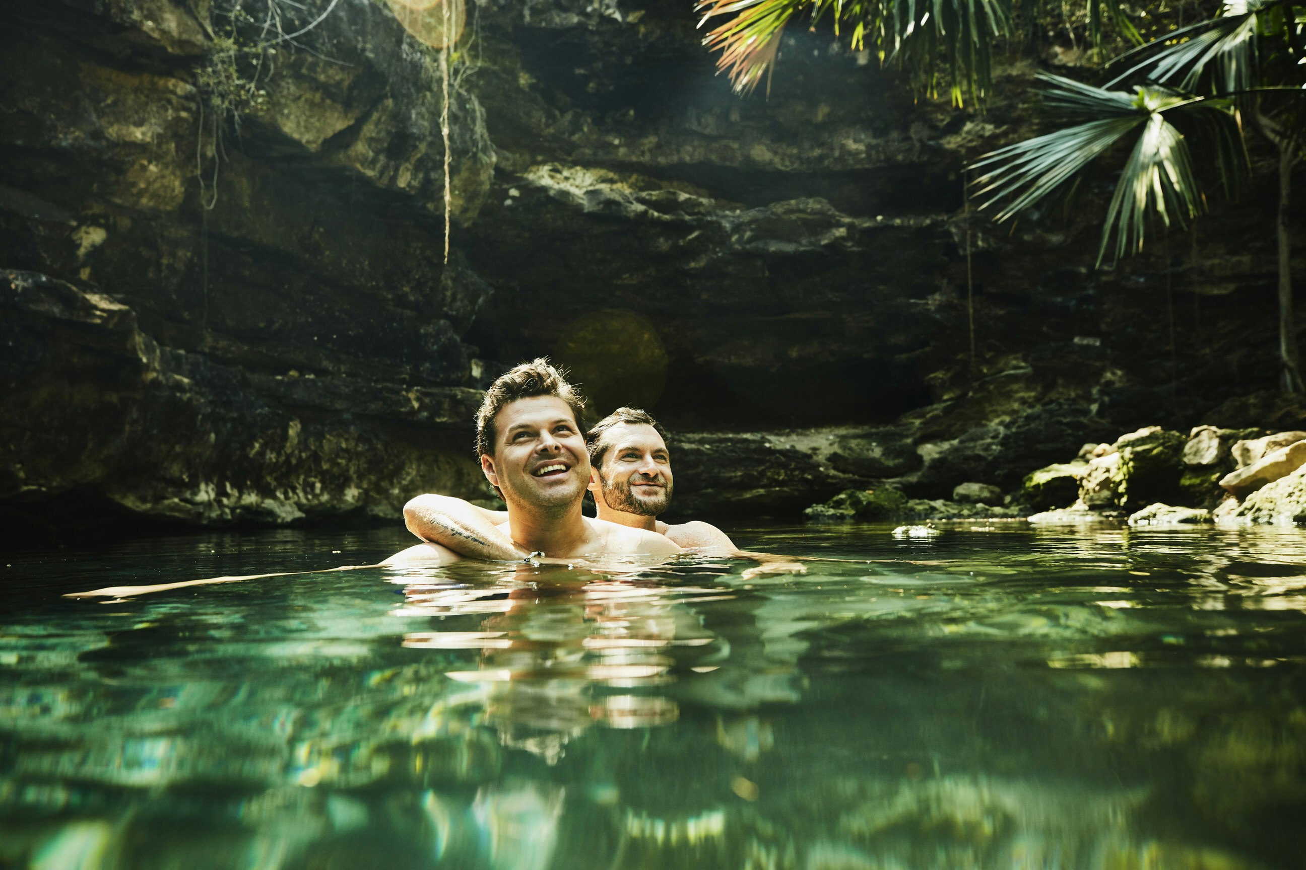 A smiling couple are swimming in the blue-green waters of a cenote. Sun shines down into the pool and highlights the two men as one is embracing the other.