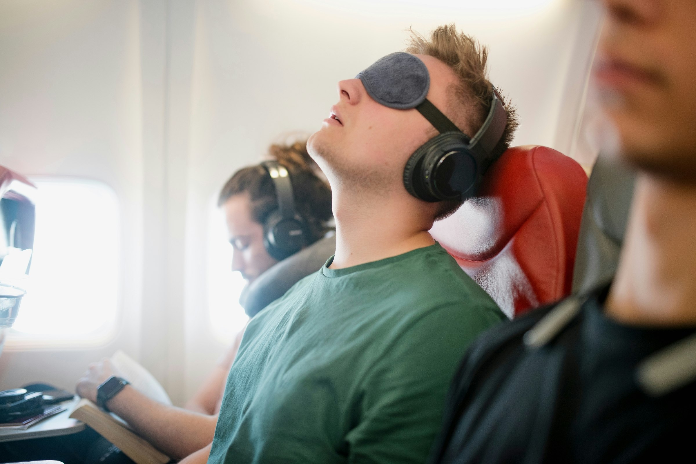 Close-up of a young man wearing wireless headphones and an eye mask sleeping on an economy flight with his head back.