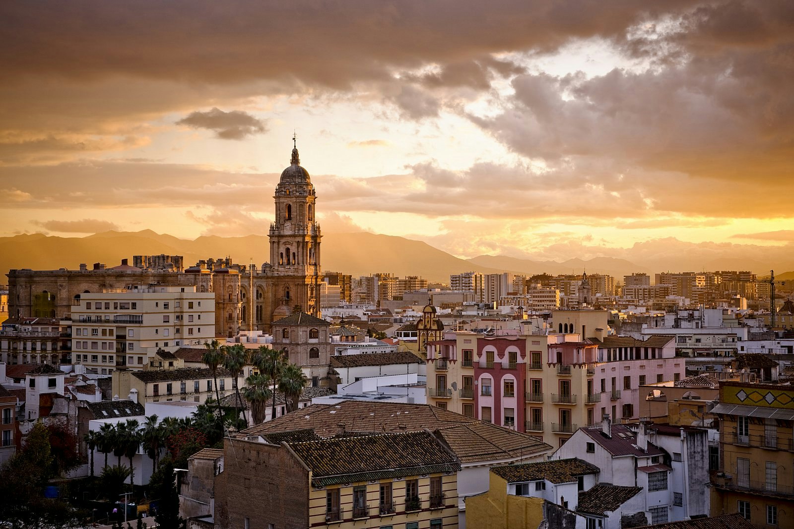 A dramatic sunset over the Malaga skyline, which has a mixture of modern and historic buildings and a church spire at its centre. 