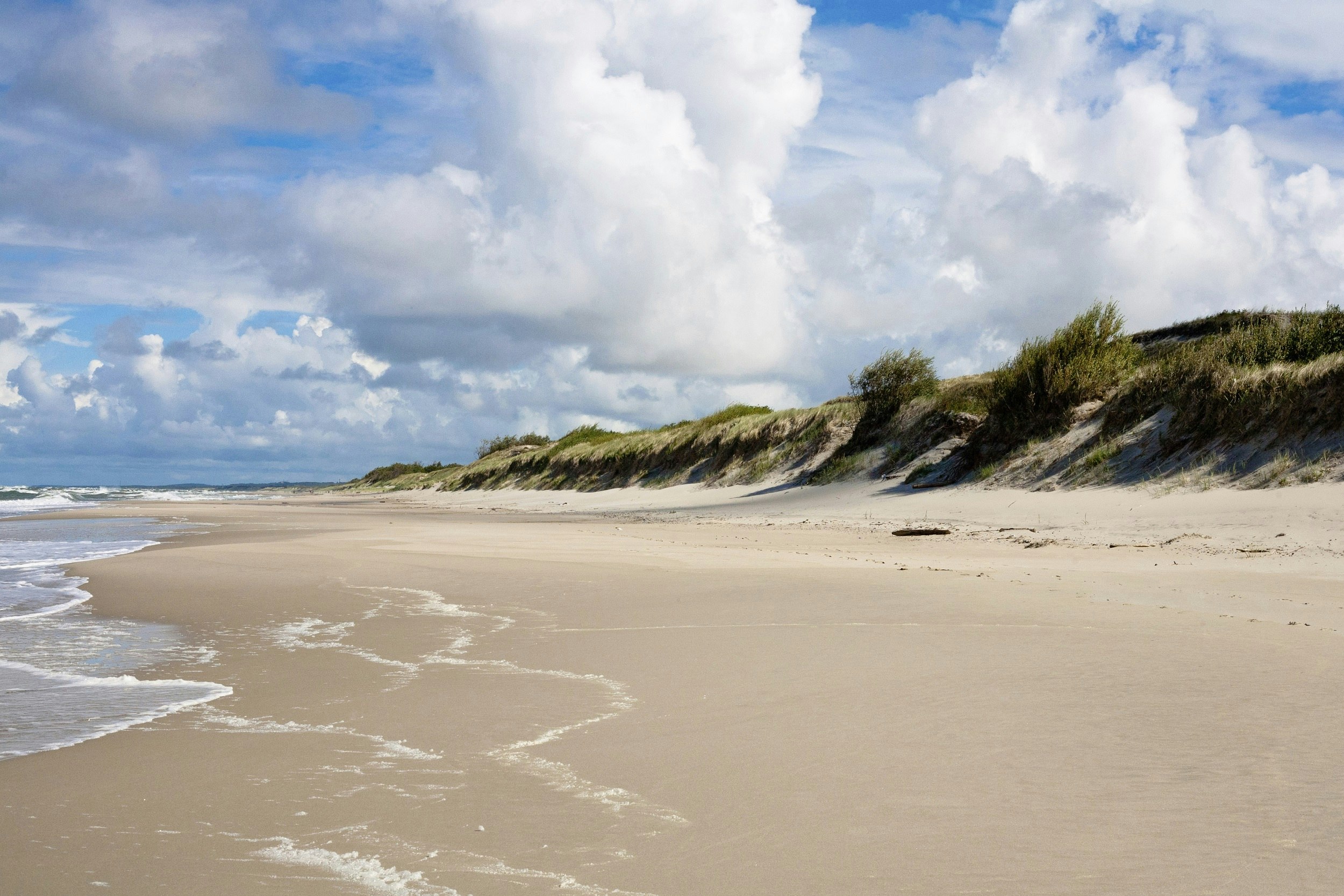 An empty beach stretches into the distance. Sand dunes covered in grass are on the right; the Baltic Sea is on the left