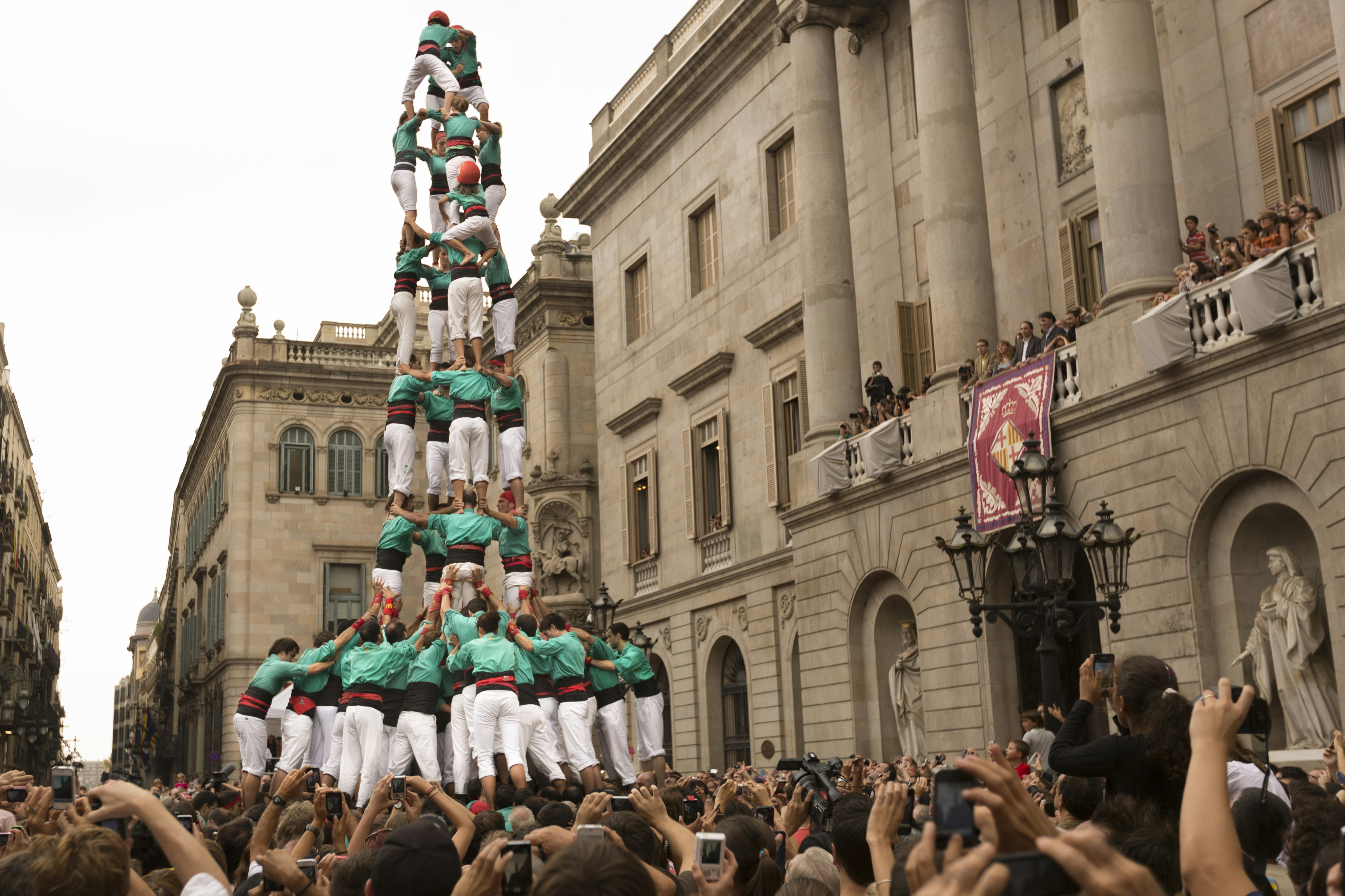 Castellers (human towers) in Barcelona's Plaça Sant Jaume during La Mercè; people wearing white trousers and green shirts are stood on each other's shoulders to form a tall tower in a square packed with people trying to photograph the spectacle.