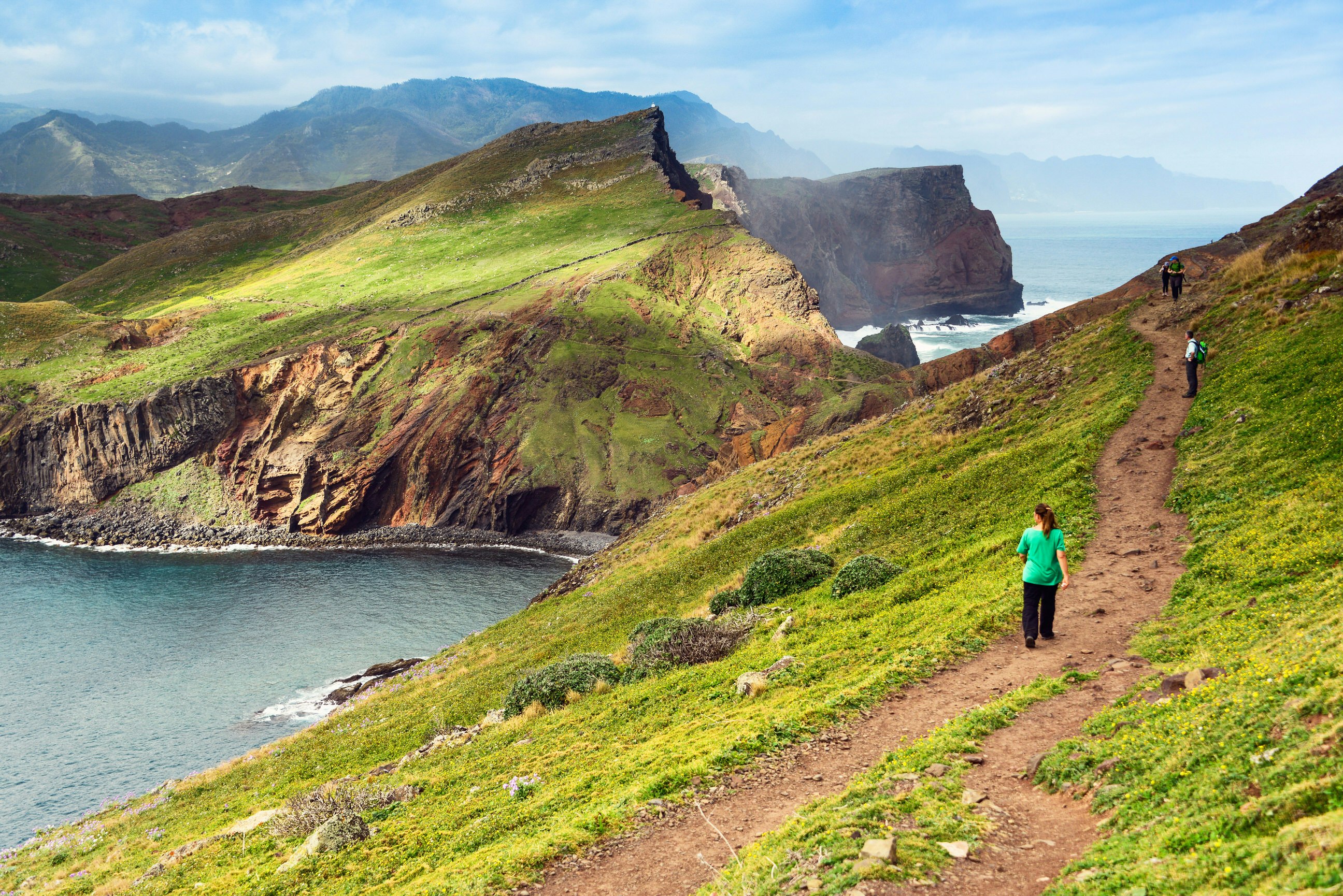 Three hikers walk along a grassy coastal trail. One is looking down towards the sea, and there are rugged mountains in the distance.