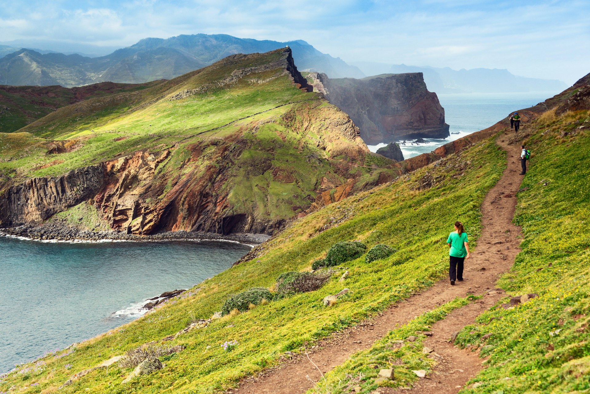 Three hikers walk along a grassy coastal trail. One is looking down towards the sea, and there are rugged mountains in the distance.