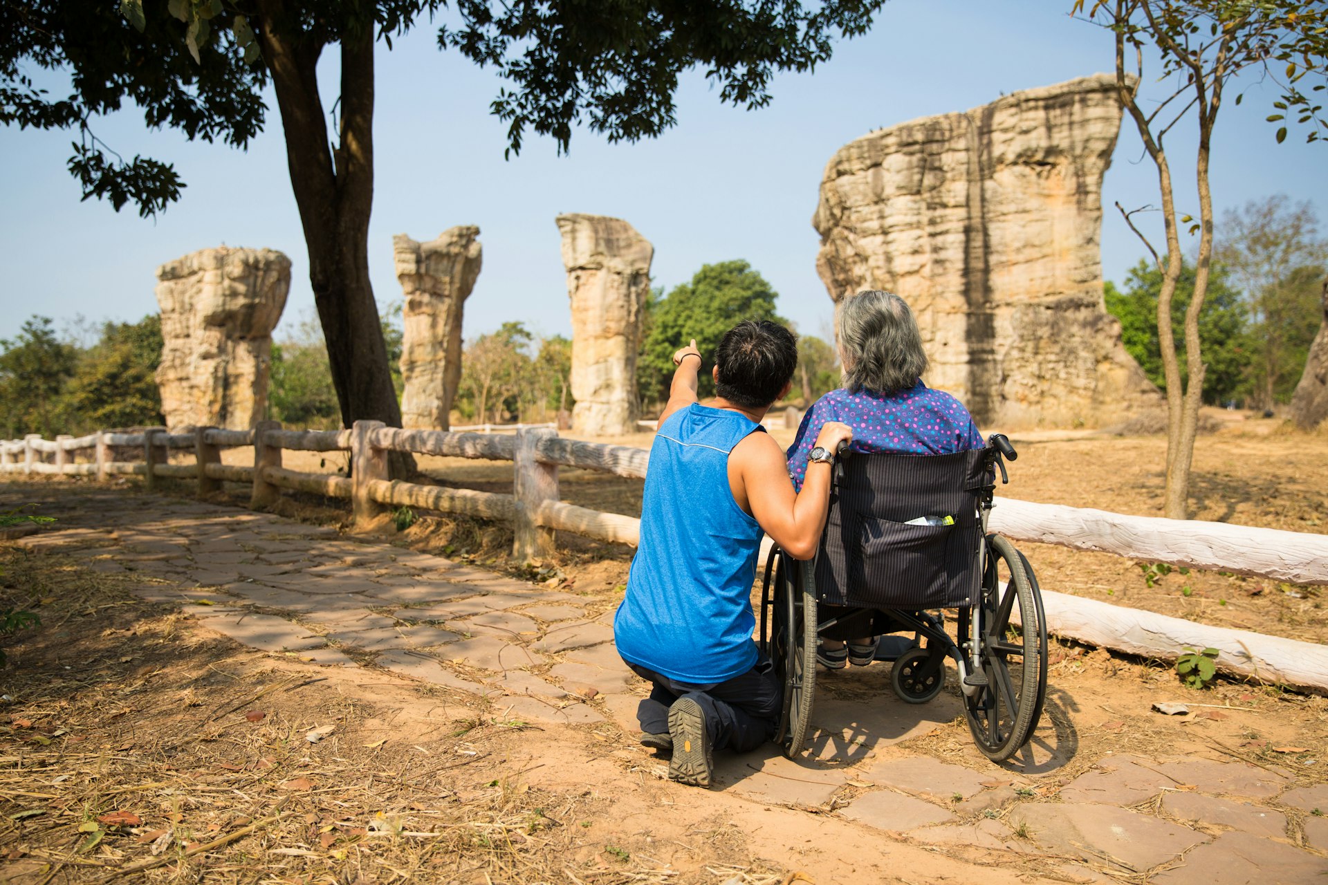 Two people look at the rock formation of Mo Hin Khao (Thailand's Stonehenge) in Chaiyaphum Province, Thailand. One is in a wheelchair and the other has crouched down to talk and point at the formation.