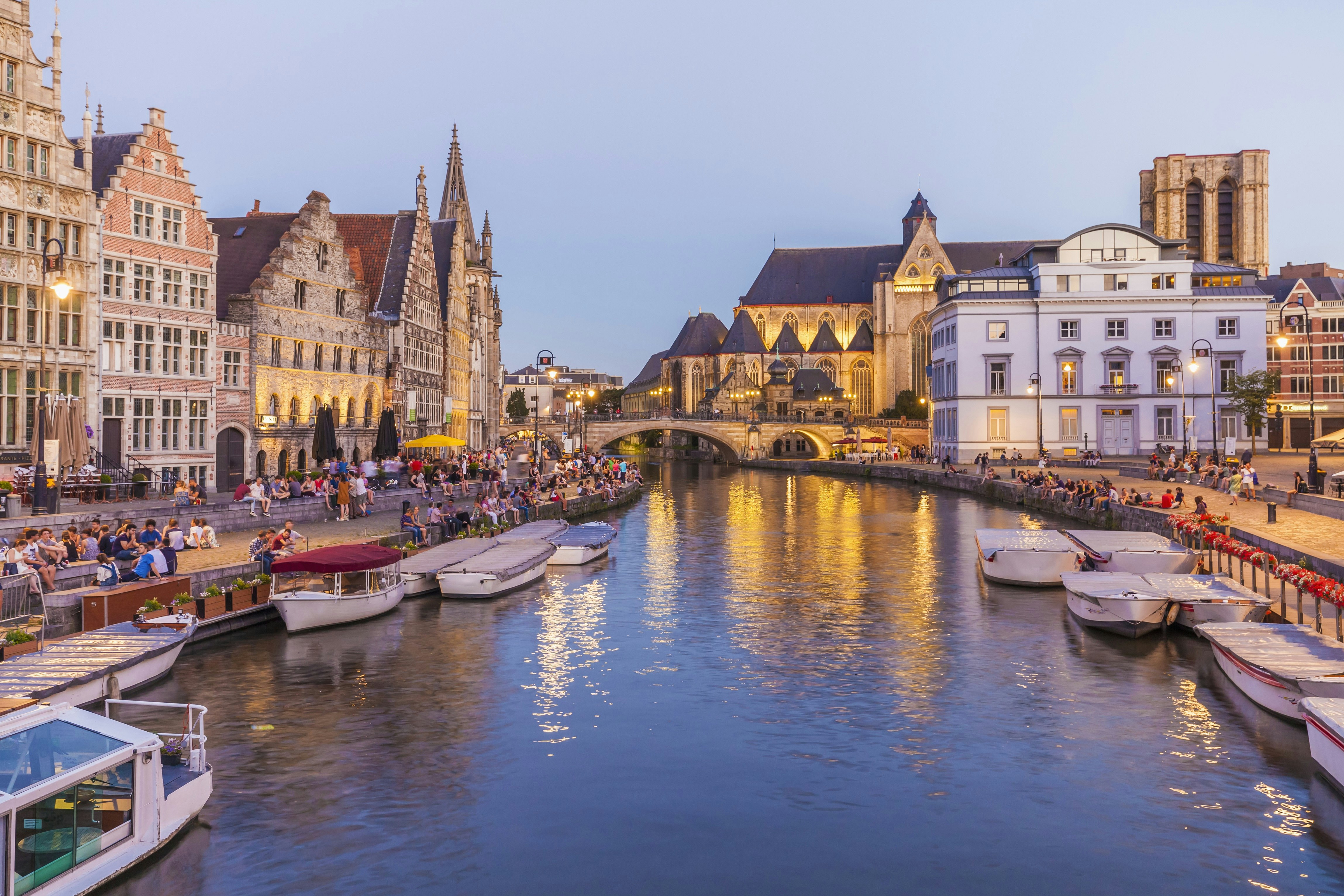 A view of Ghent's old town at dusk, with historic houses and churches spread around the River Leie. Several boats are moored on the river, while groups of people sit on its banks.