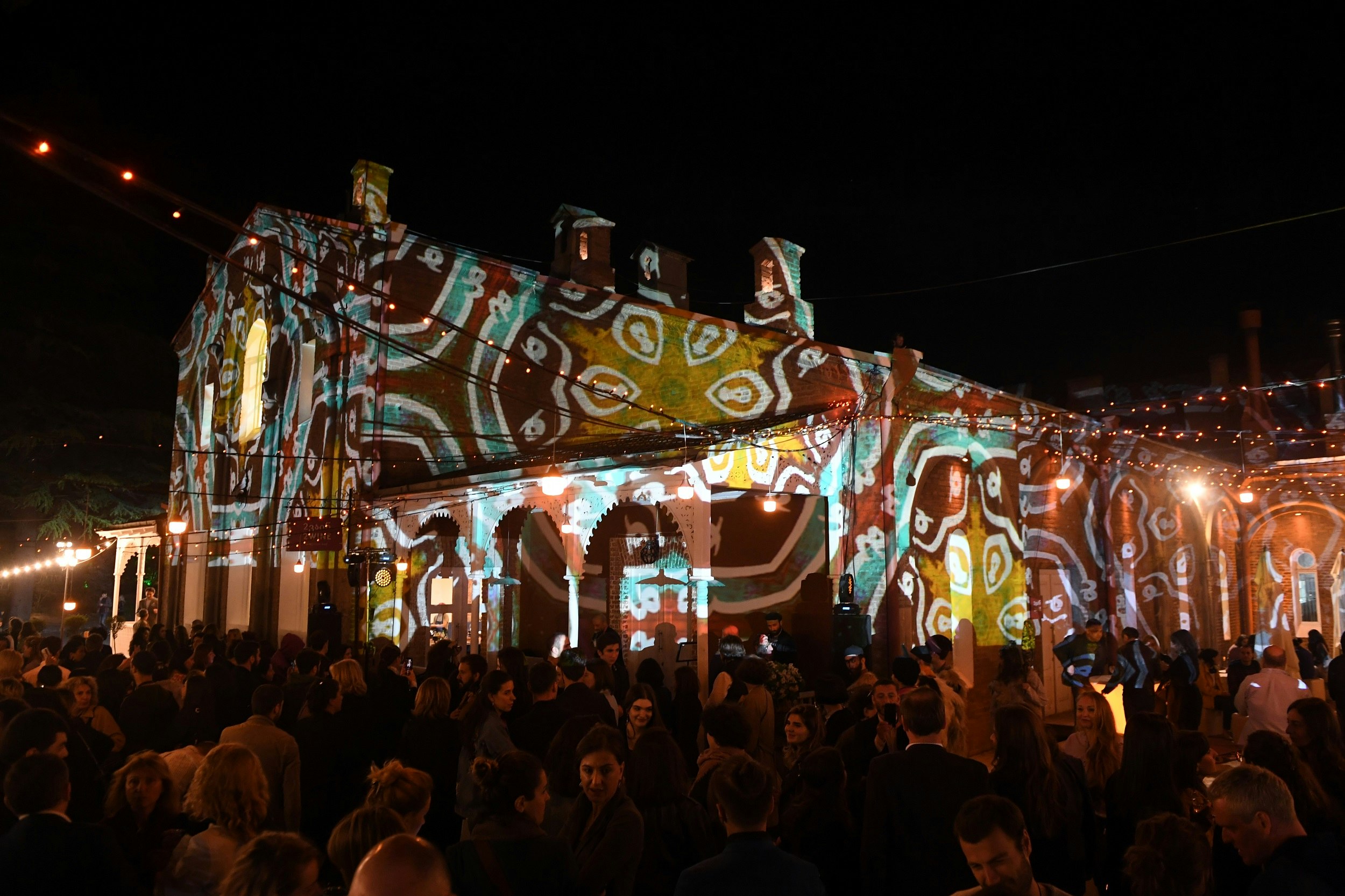 Patterns are projected by lights onto a building. The area is crowded with people socialising