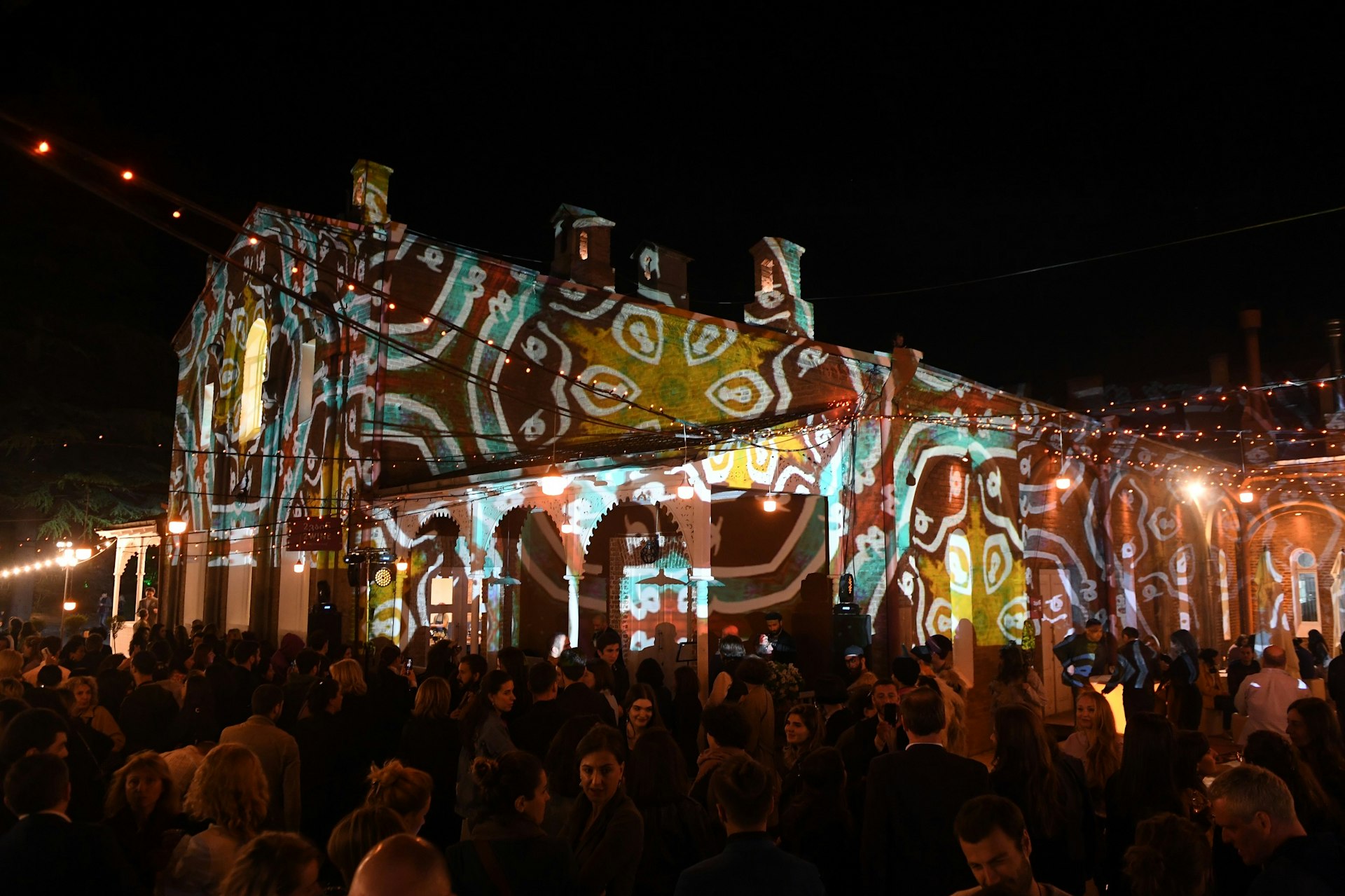 Patterns are projected by lights onto a building. The area is crowded with people socialising