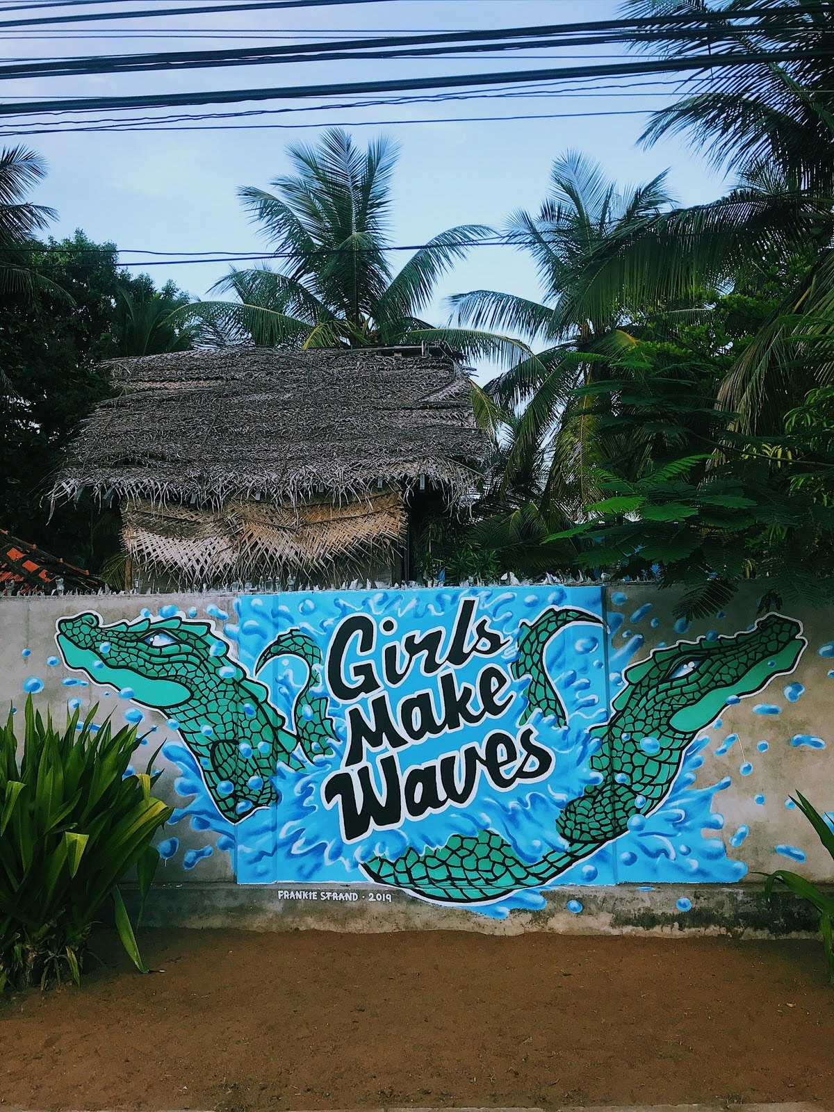A mural painted on a grey concrete wall depicting two cartoon-style crocodiles swimming out of a splash of blue water with the words 'girls make waves' in the centre of the image