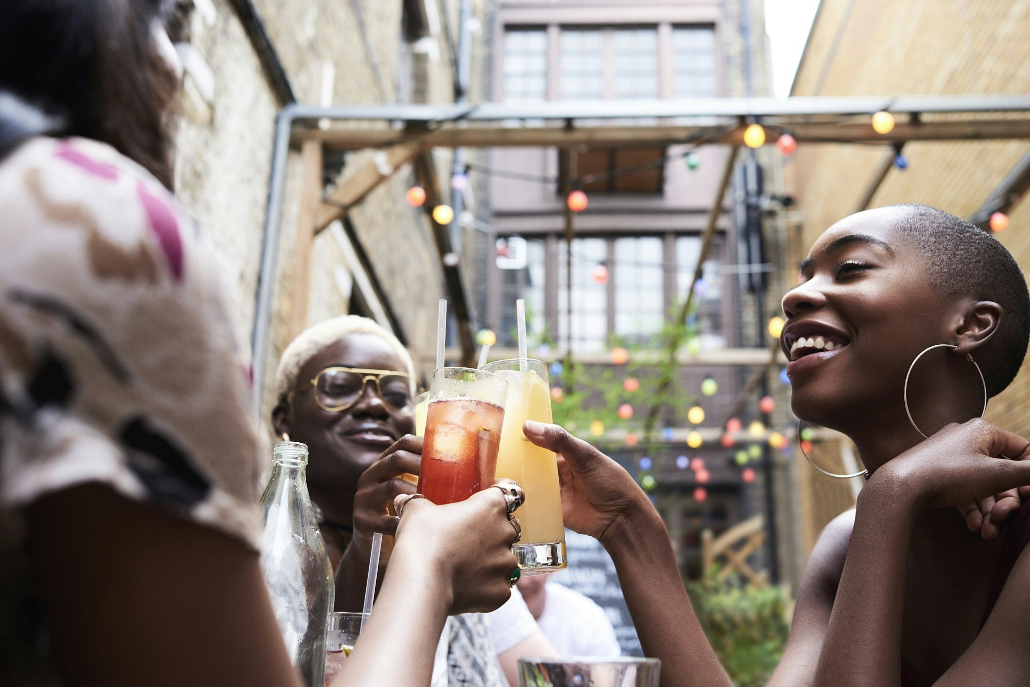 Tre flickor skålar med cocktails i en utomhusbar med färgade lampor ovanför.