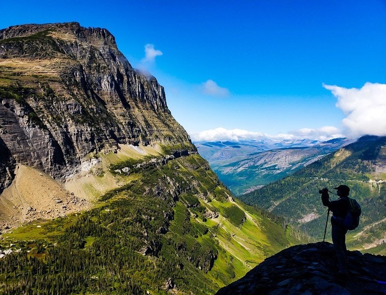 A person taking a photo is silhouetted against a green valley with large mountains