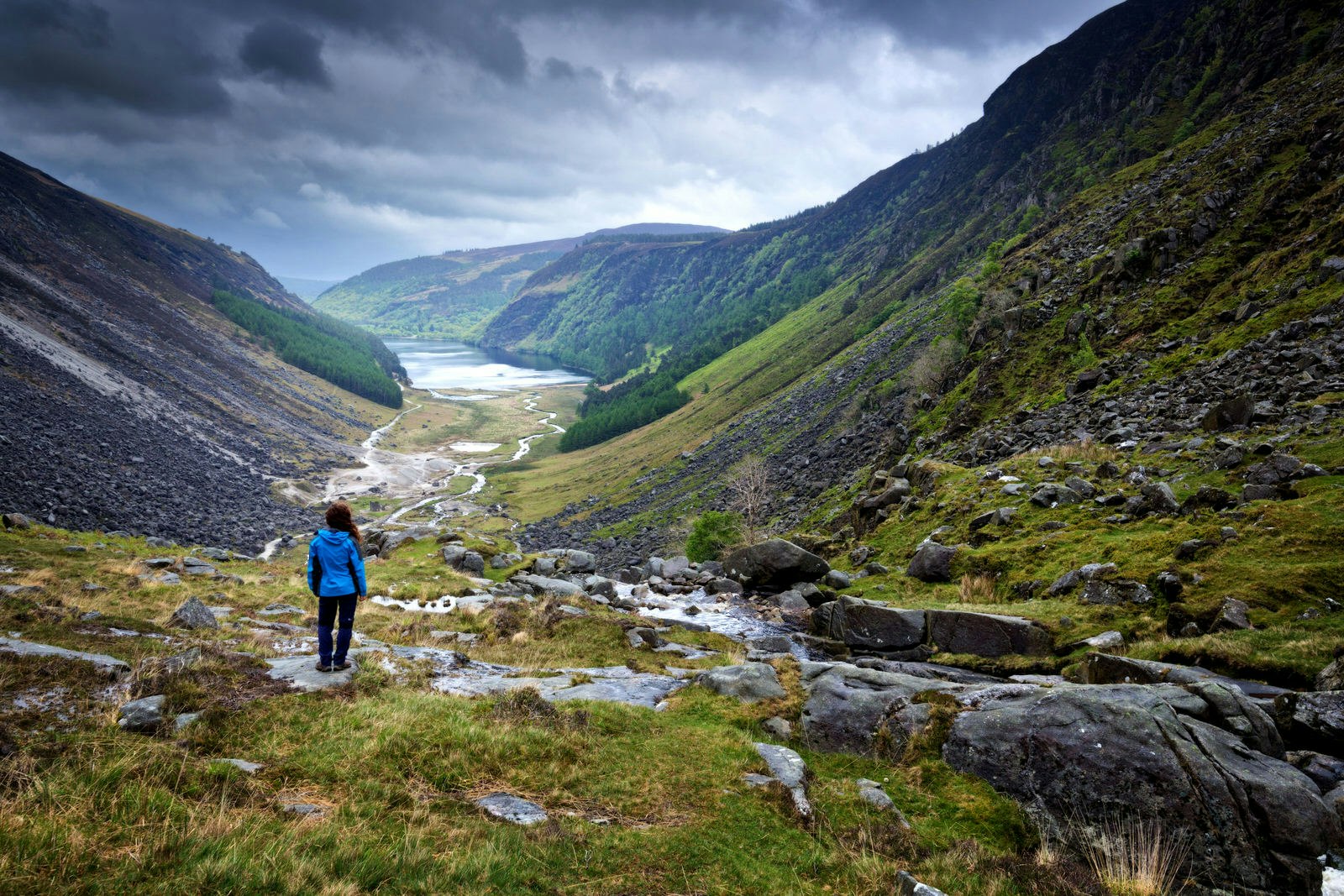 A person in a blue coat stands looking at Glendalough, a valley carved by a glacier.