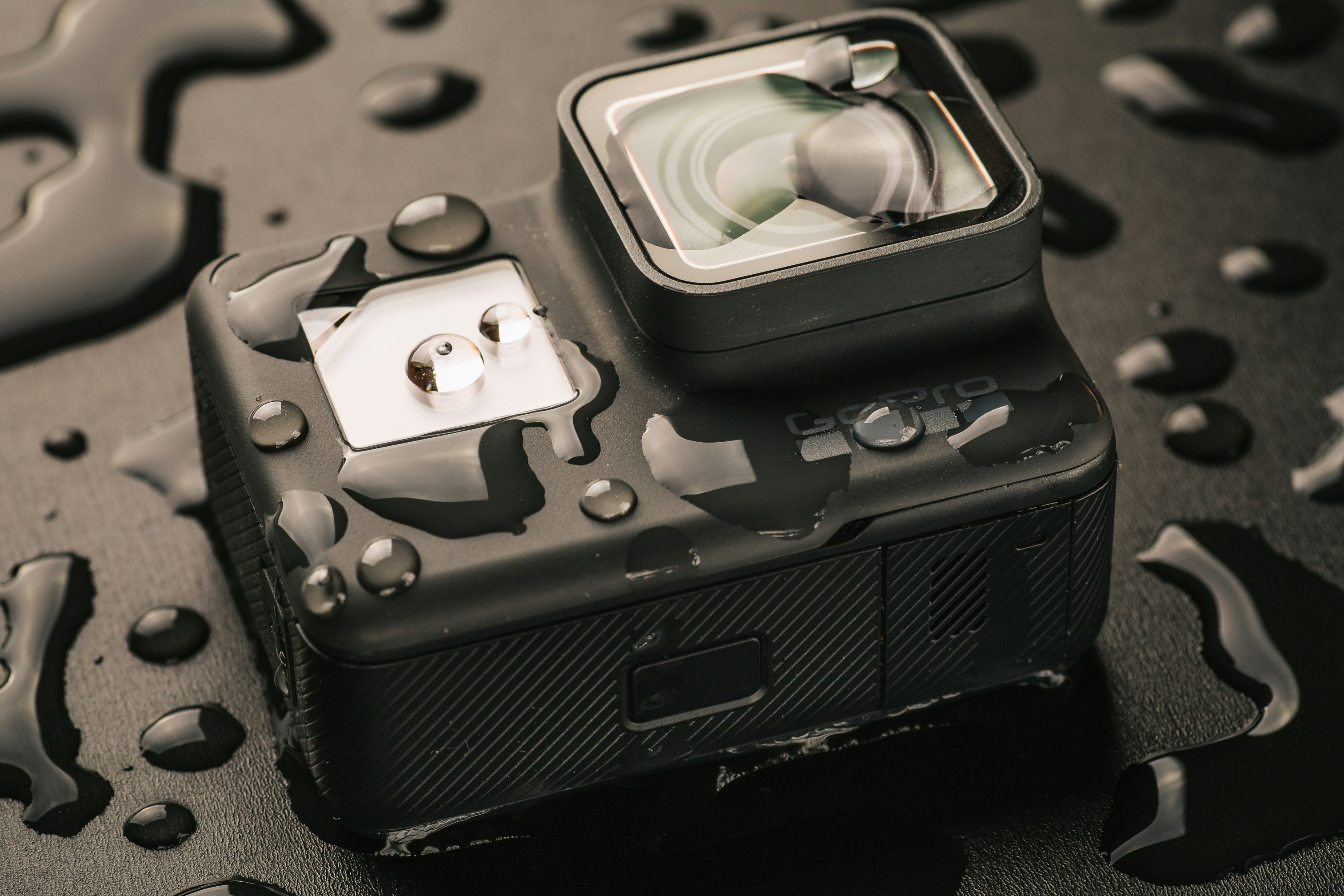 A GoPro Camera is on a black surface, covered in globules of water. 