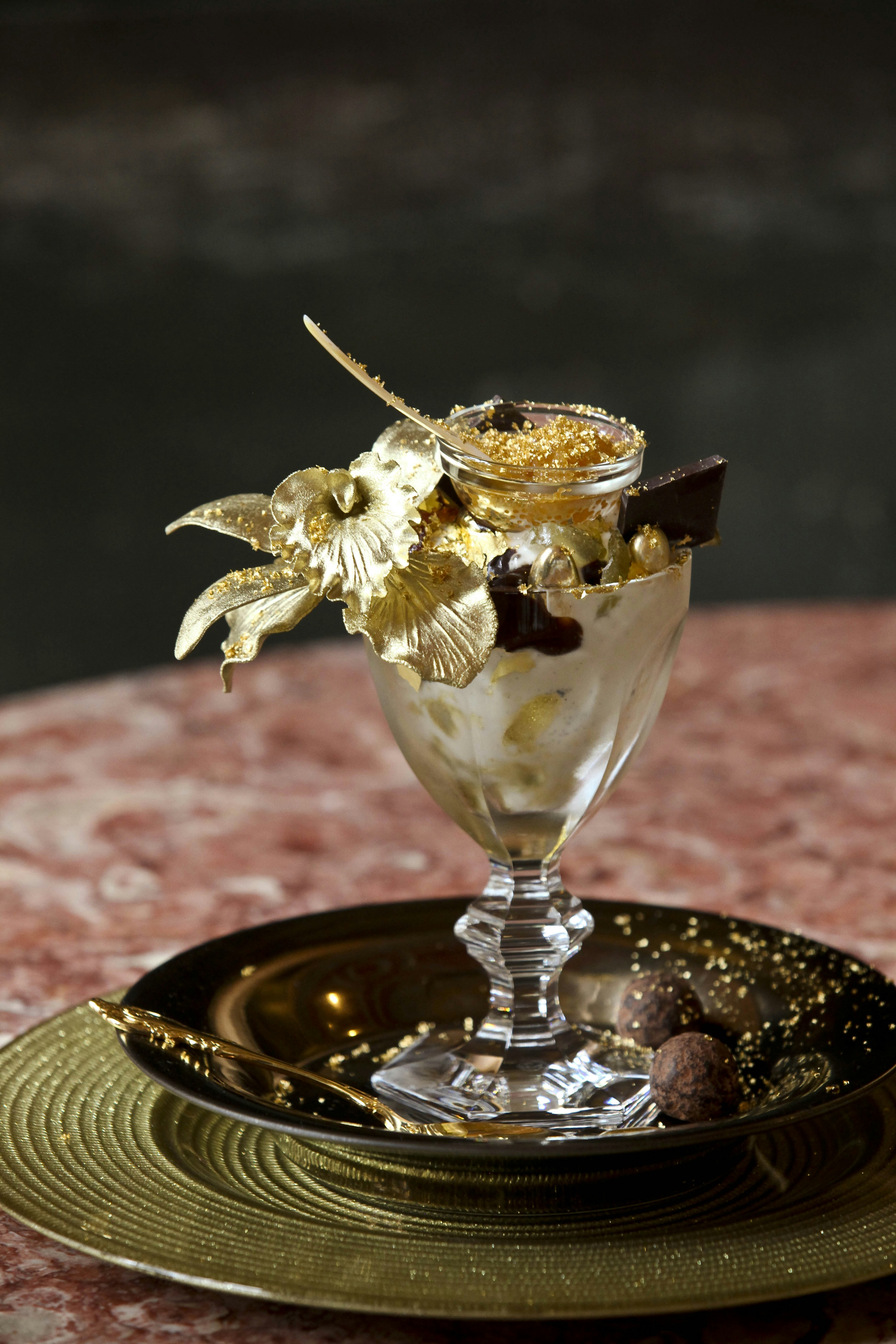A small container of gold-dusted caviar rests atop an ice cream sundae of vanilla ice cream filled with Chuao chocolate, golden pieces of candy and garnished with a sugar forged orchid. There is a golden spoon resting at the base of the $350 Baccarat Harcourt crystal goblet
