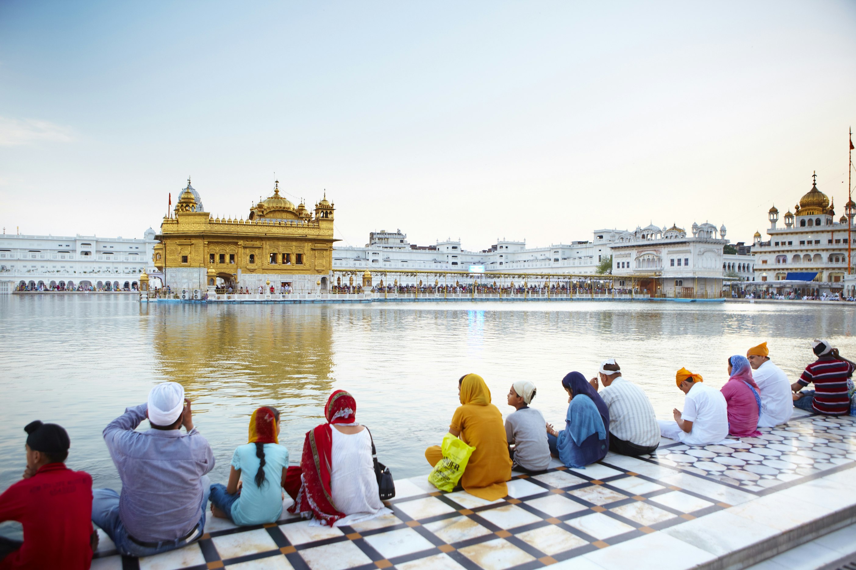 A row of people are sitting at the edge of Amrit Sarovar lake with their backs to the camera, looking towards Amritsar's Golden Temple.