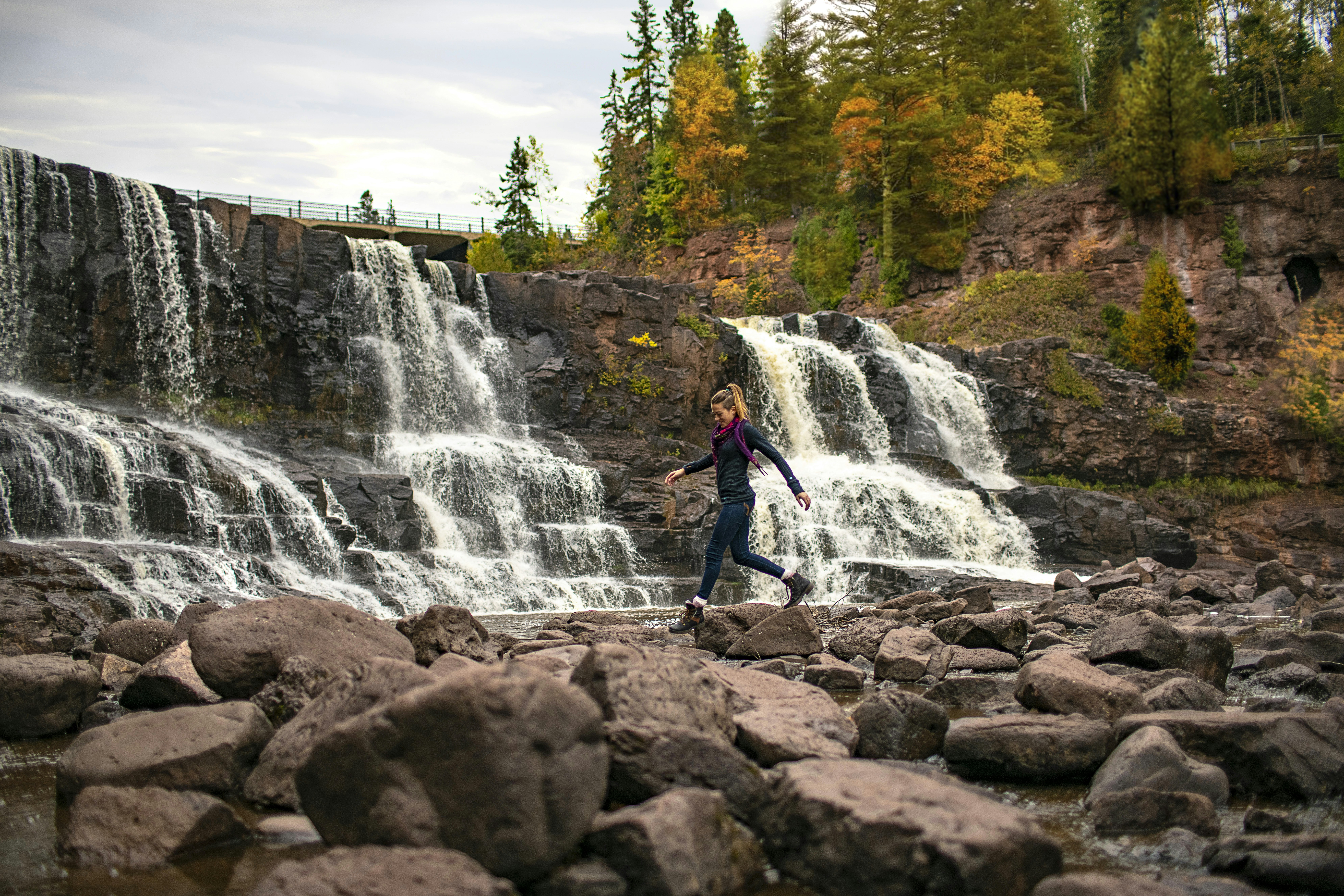 A woman hops across rocks in front of a waterfall