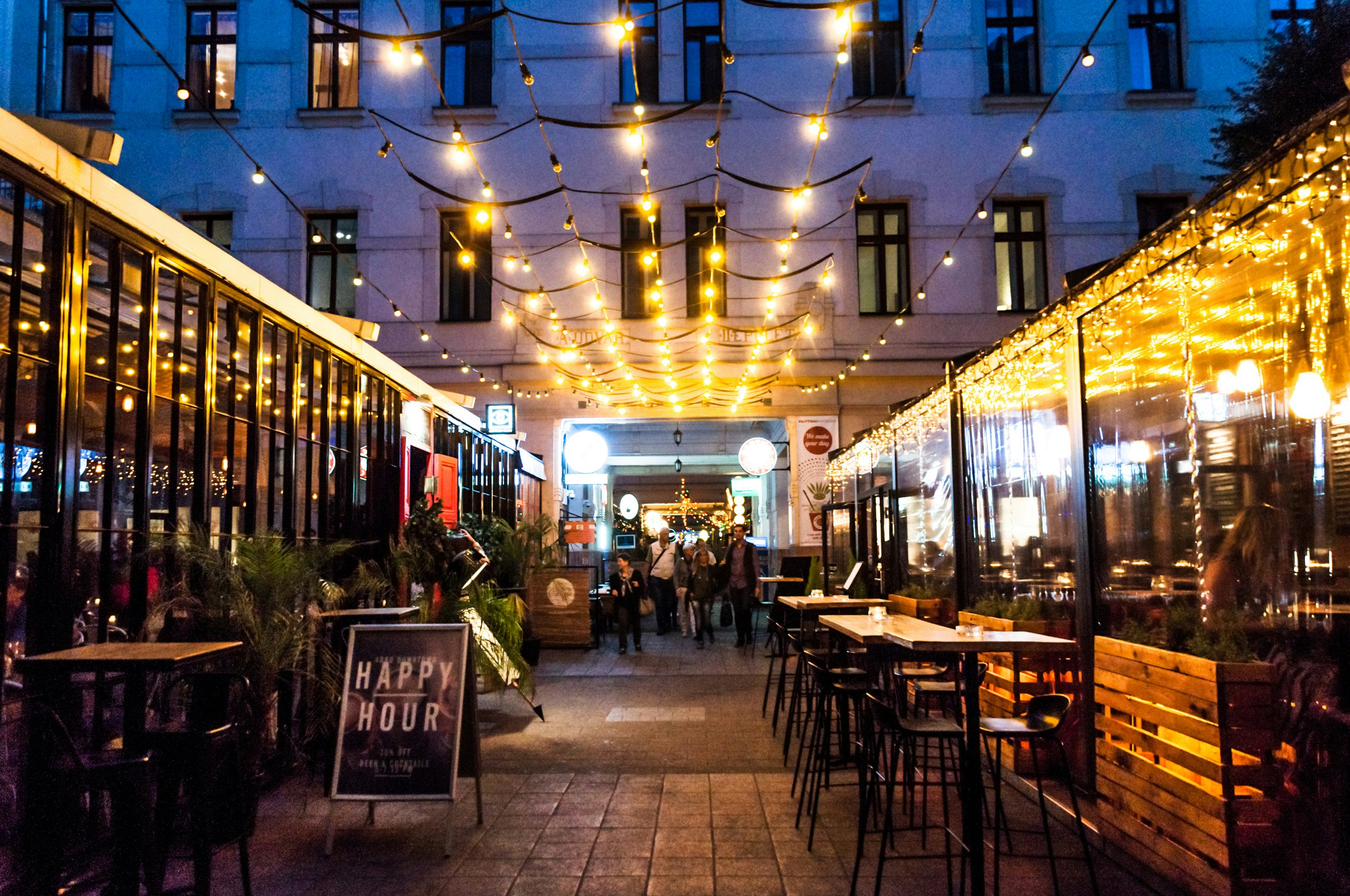 A passageway between buildings lined with string lights. A bar on the left has a Happy Hour sign outside it. Tables and stools line the passage.