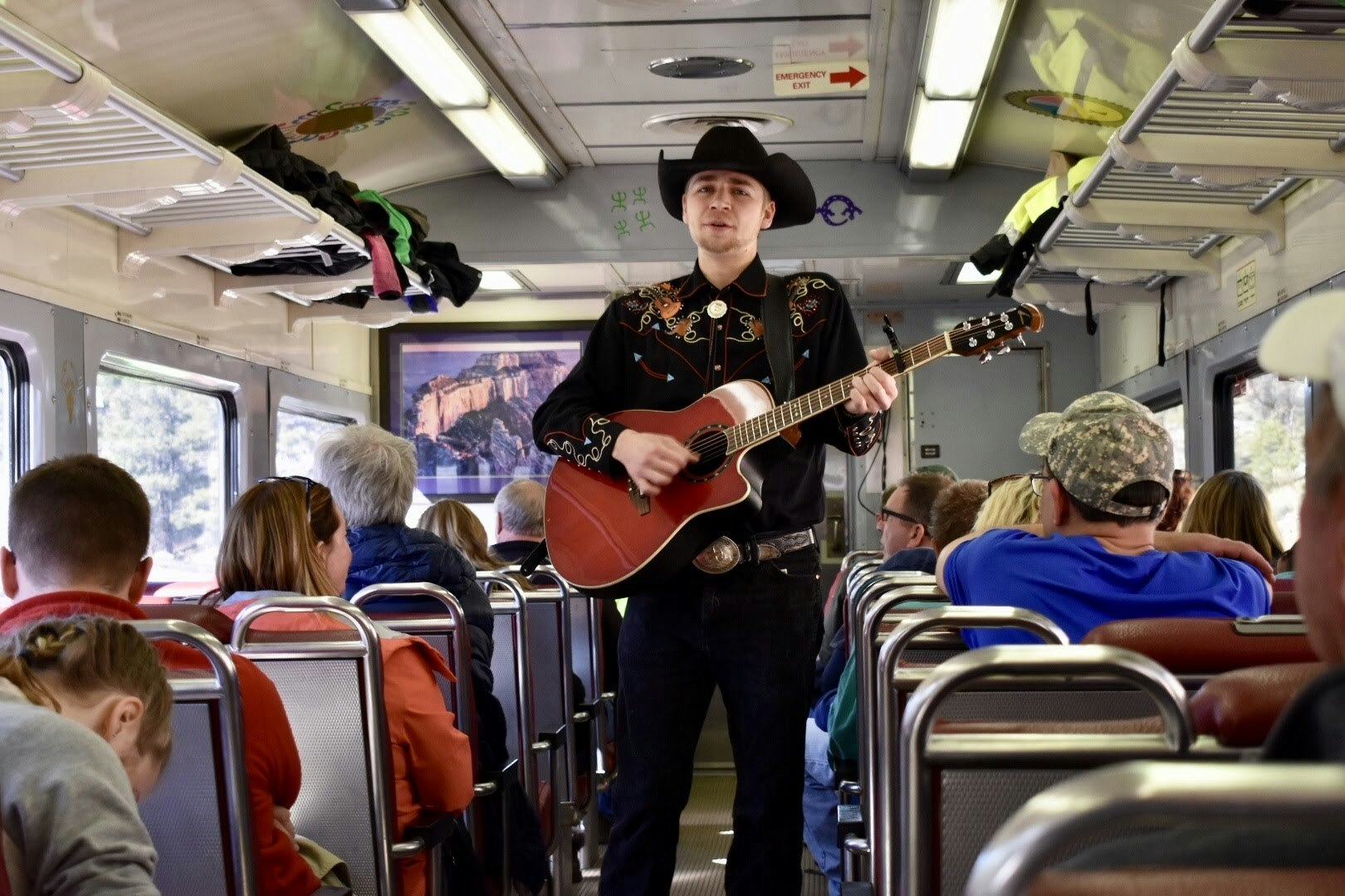A young man in an embroidered shirt and black cowboy hat strums a guitar and sings in a train car