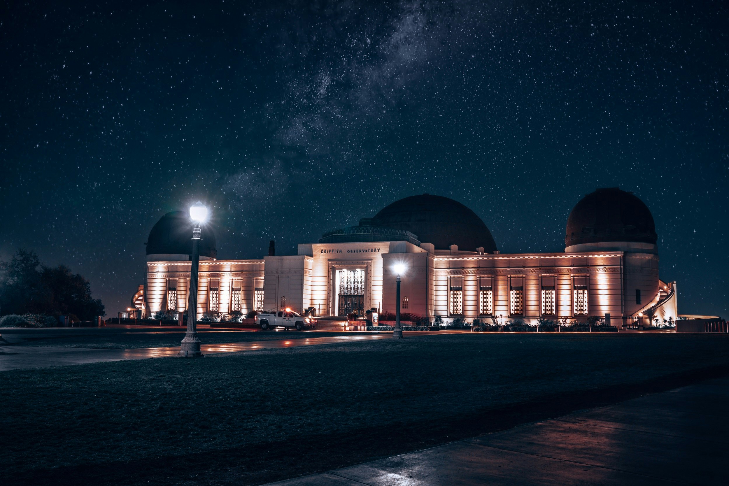 The art deco facade of the Griffith Observatory is let up at night, with the Milky Way splashed across the night sky behind it.