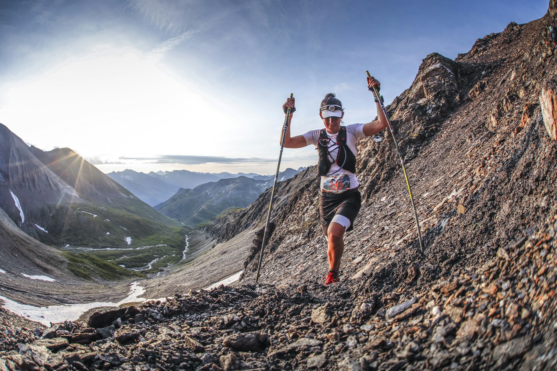 A female runner with two long poles ascends a scree slope towards the camera on the side of a dramatic valley; in the background the valley descends back towards the lowering sun.