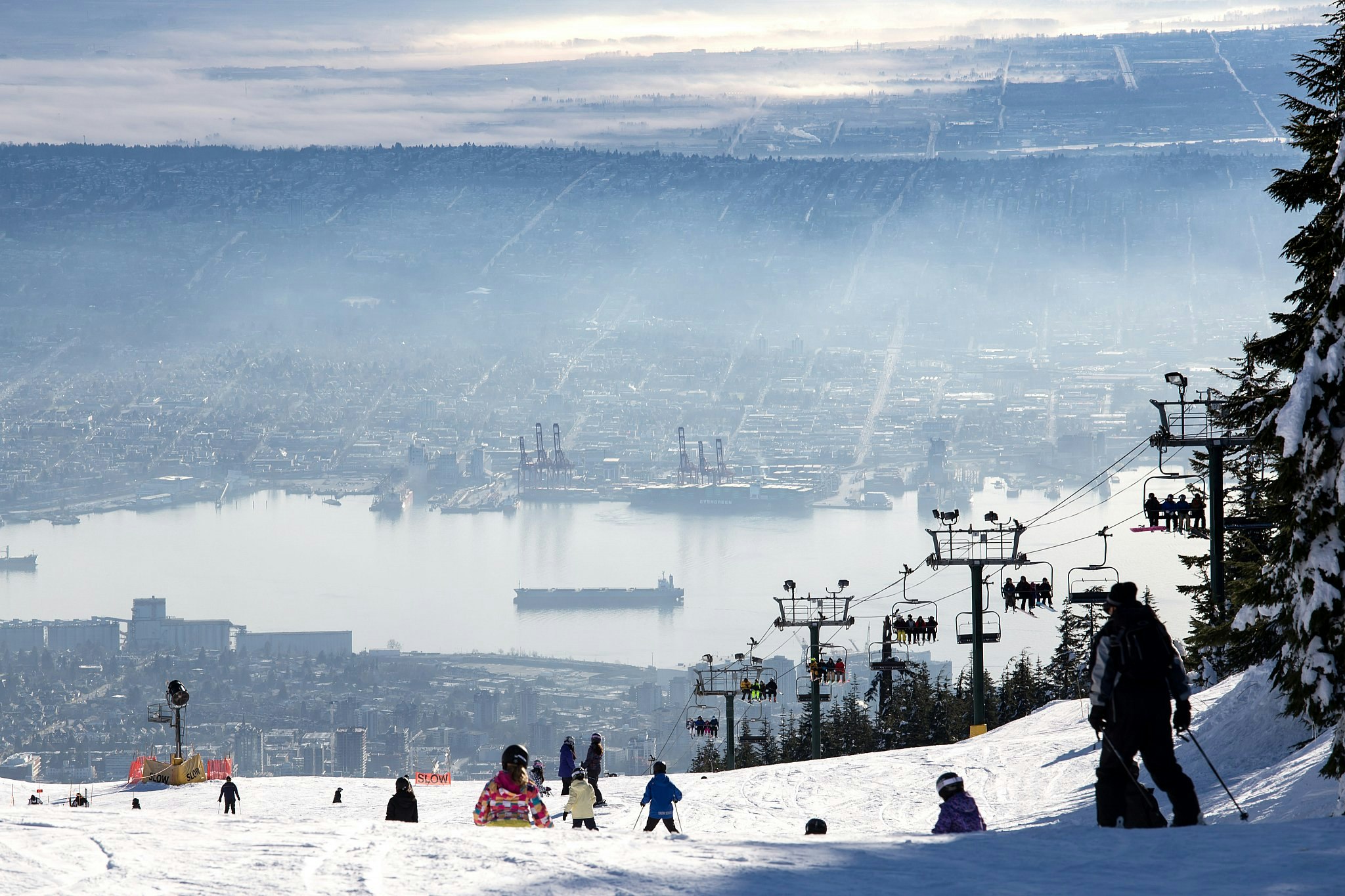 Skiers on a ski slope at the top of Grouse Mountain in Vancouver, next to trees and a ski lift; below is the city skyline, bisected by a wide harbour.