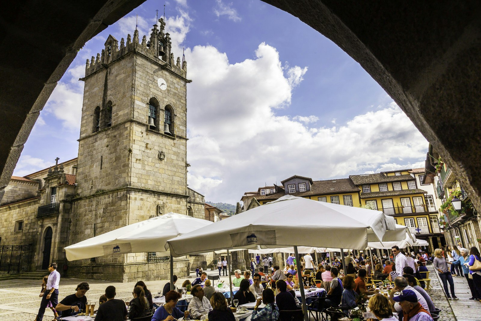 An archway view of Guimarães city square, with outdoor restaurants and tourists seen socialising; a large church rises in one corner of the square. 